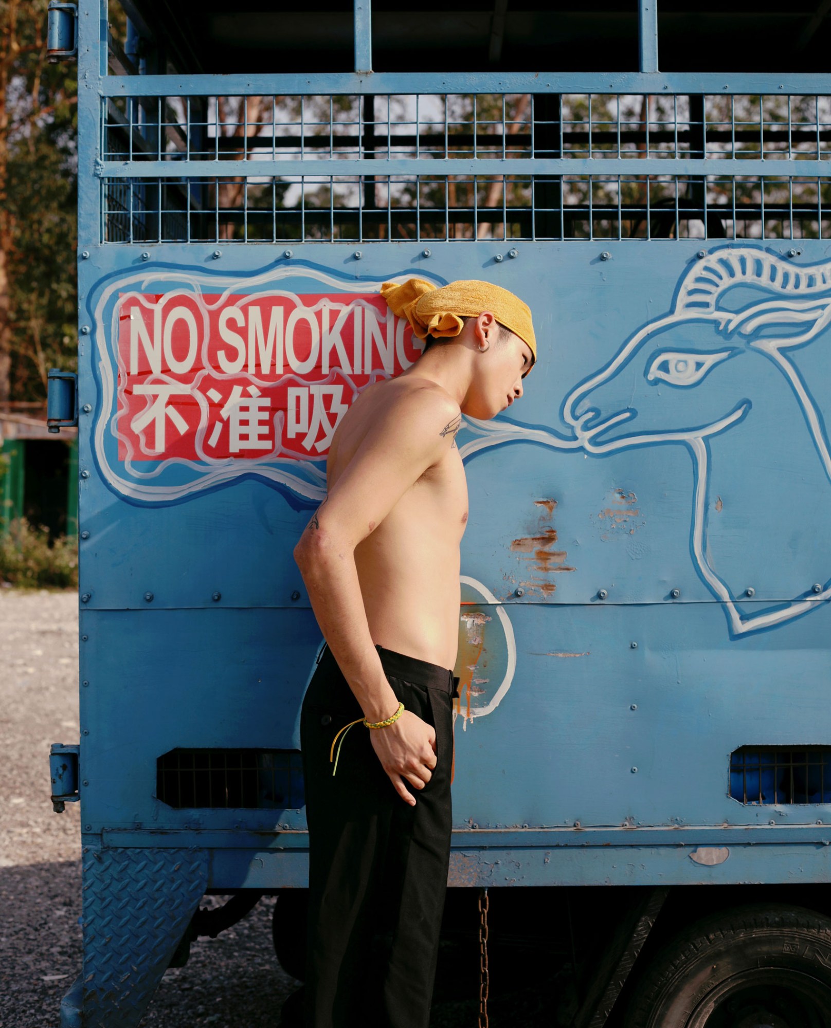 a young man wearing black trousers and a yellow headscarf leans sideways against an old blue truck