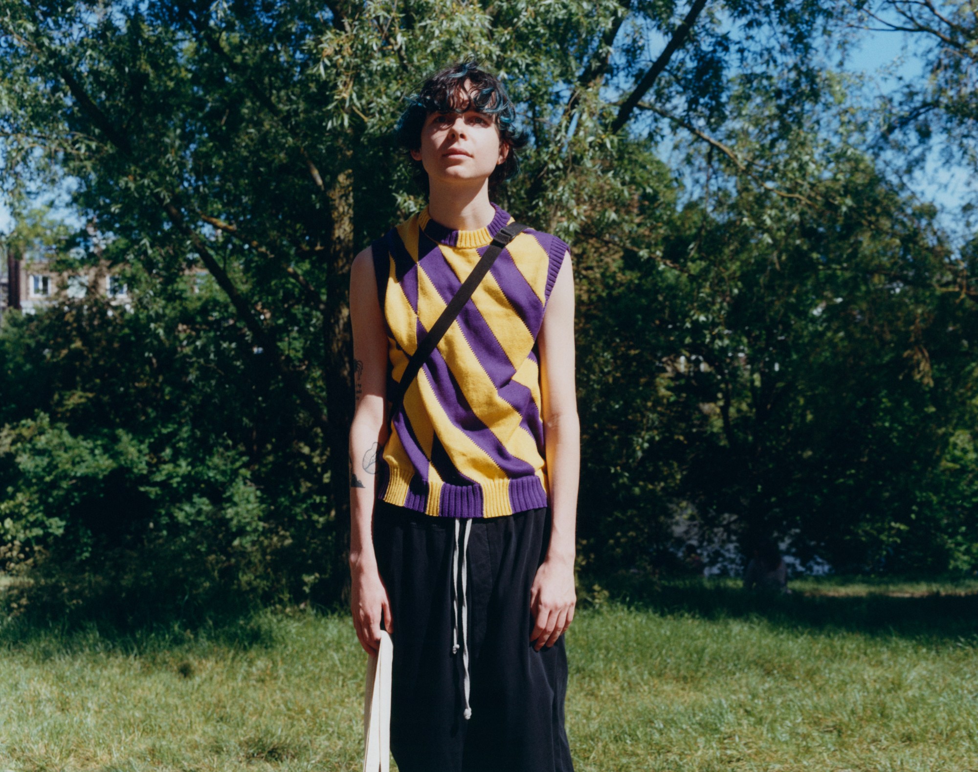 a boy with blue hair, wearing a purple and yellow knitted tank top, stands in front of trees