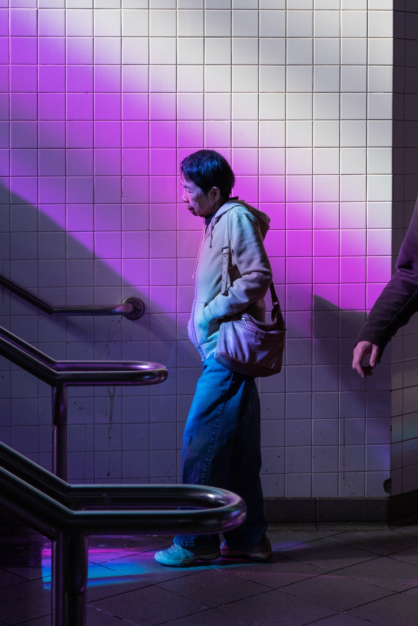 a person wearing jeans and a white hoodie walks in front of a white tiled wall of a new york subway station