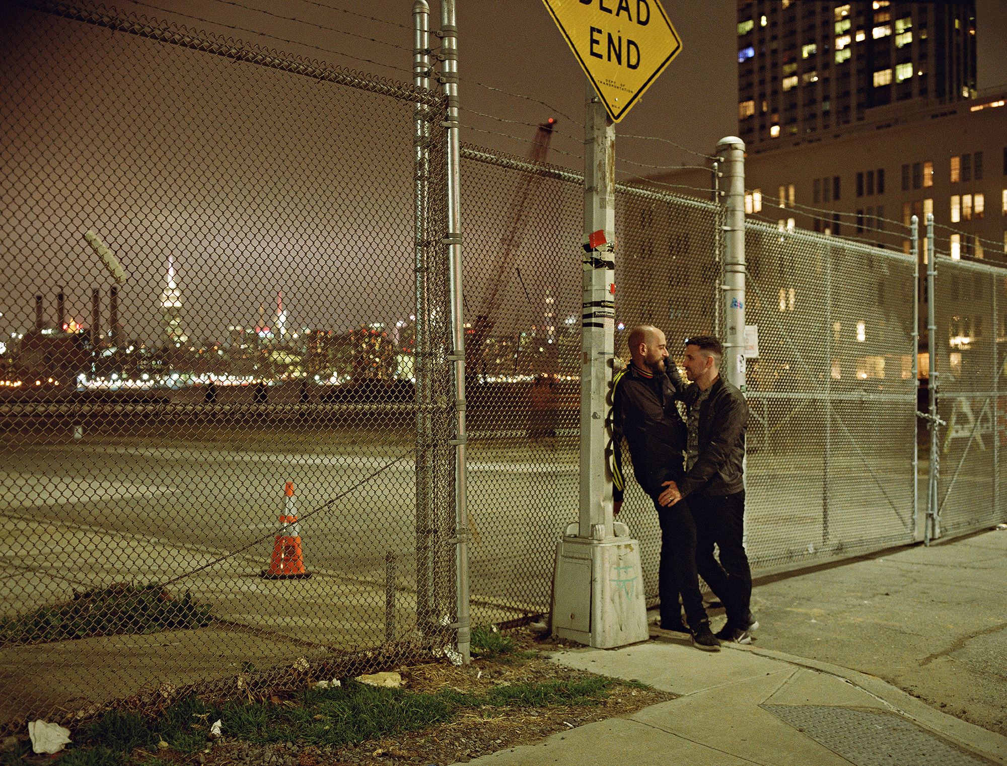 two men lean against a lampppost next to a fence with new york city behind it