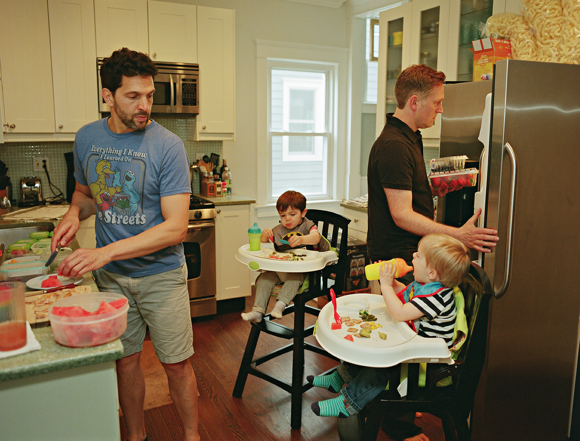 two fathers prepare breakfast for their to children in highchairs in the kitchen