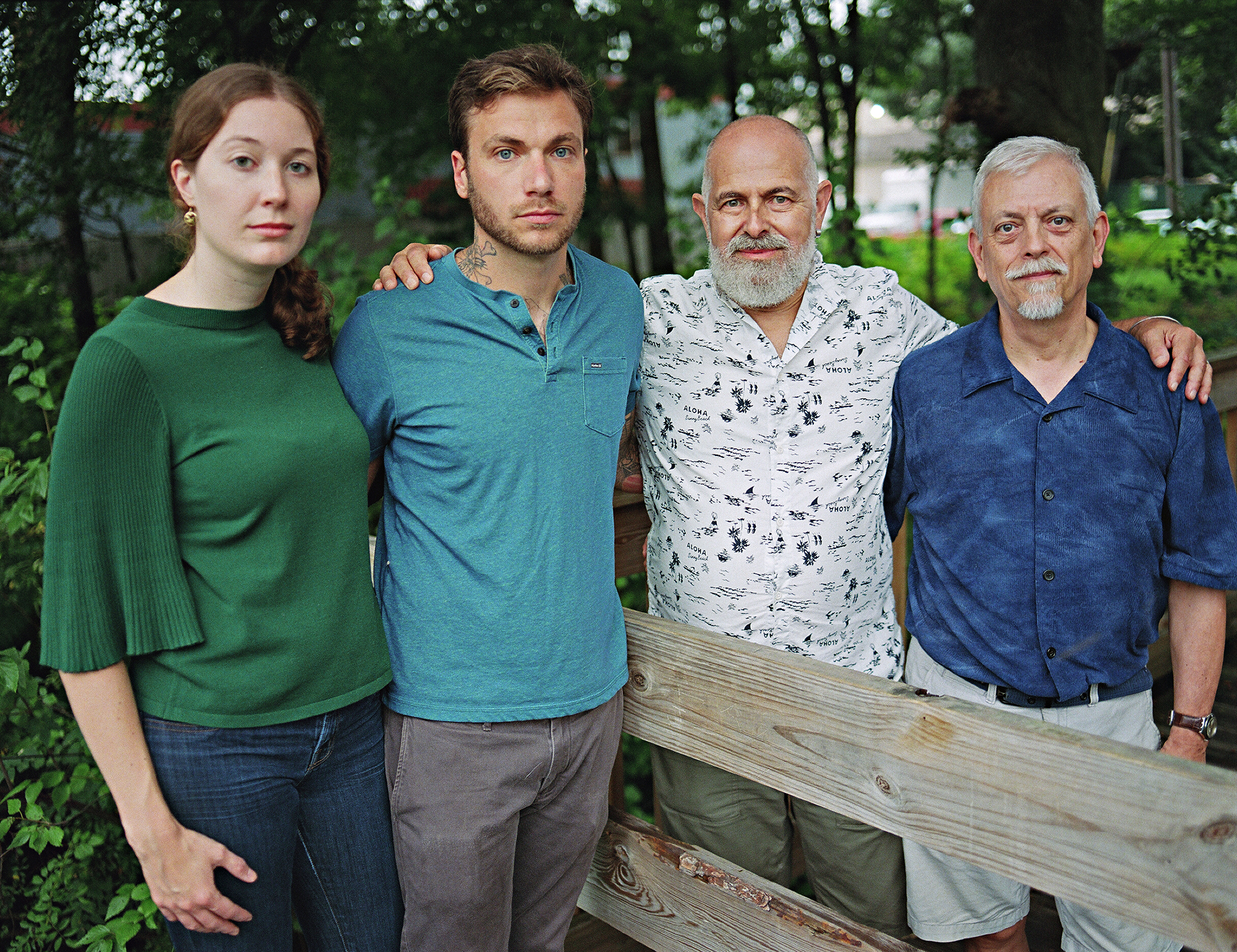 two gay men look into the camera posing with their son and his partner, outdoors and with a fence separating them