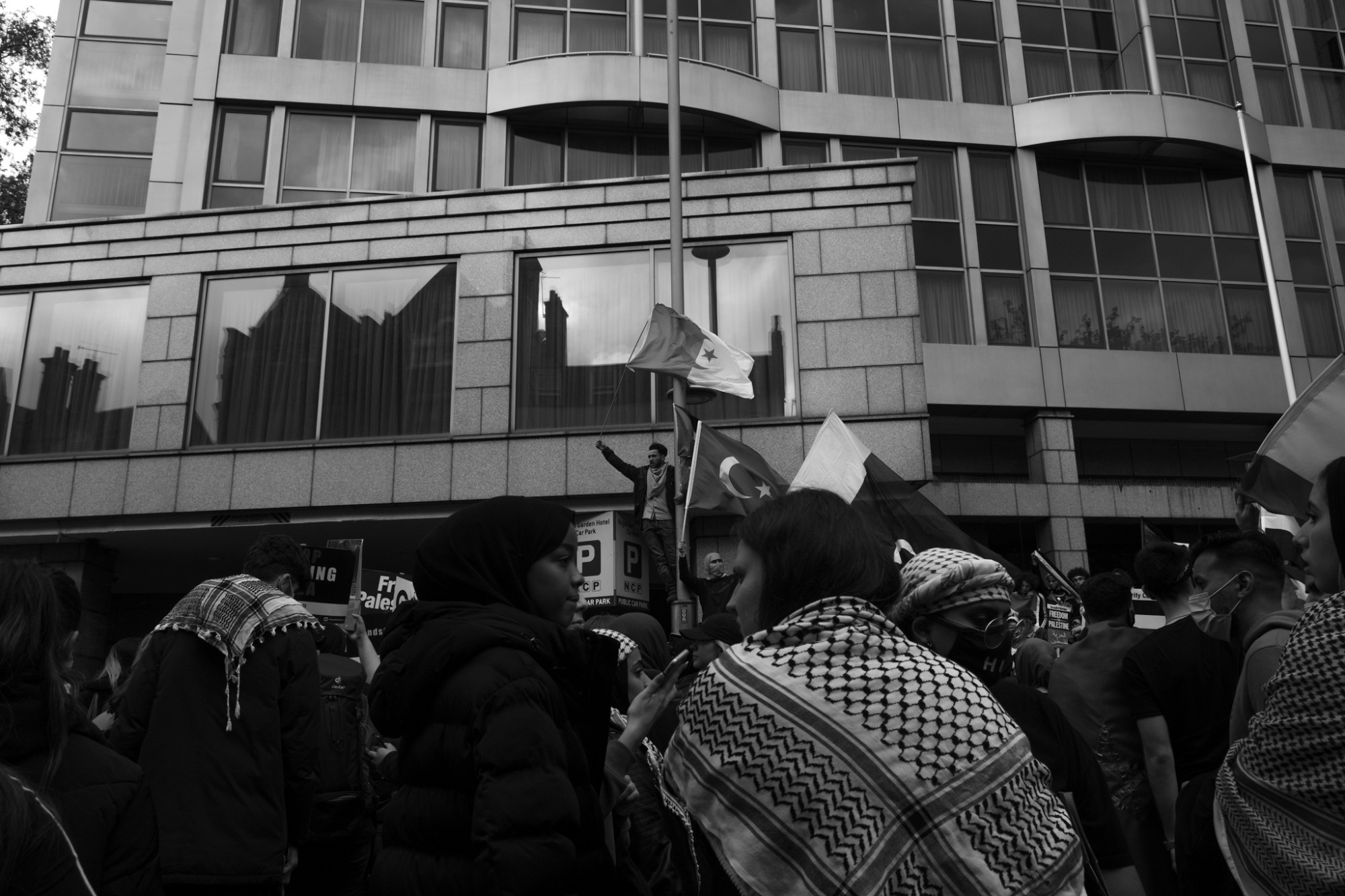 a man waving a palestinian flat stands with his fist raised above a marching crowd. two young women look at each other in the foreground.