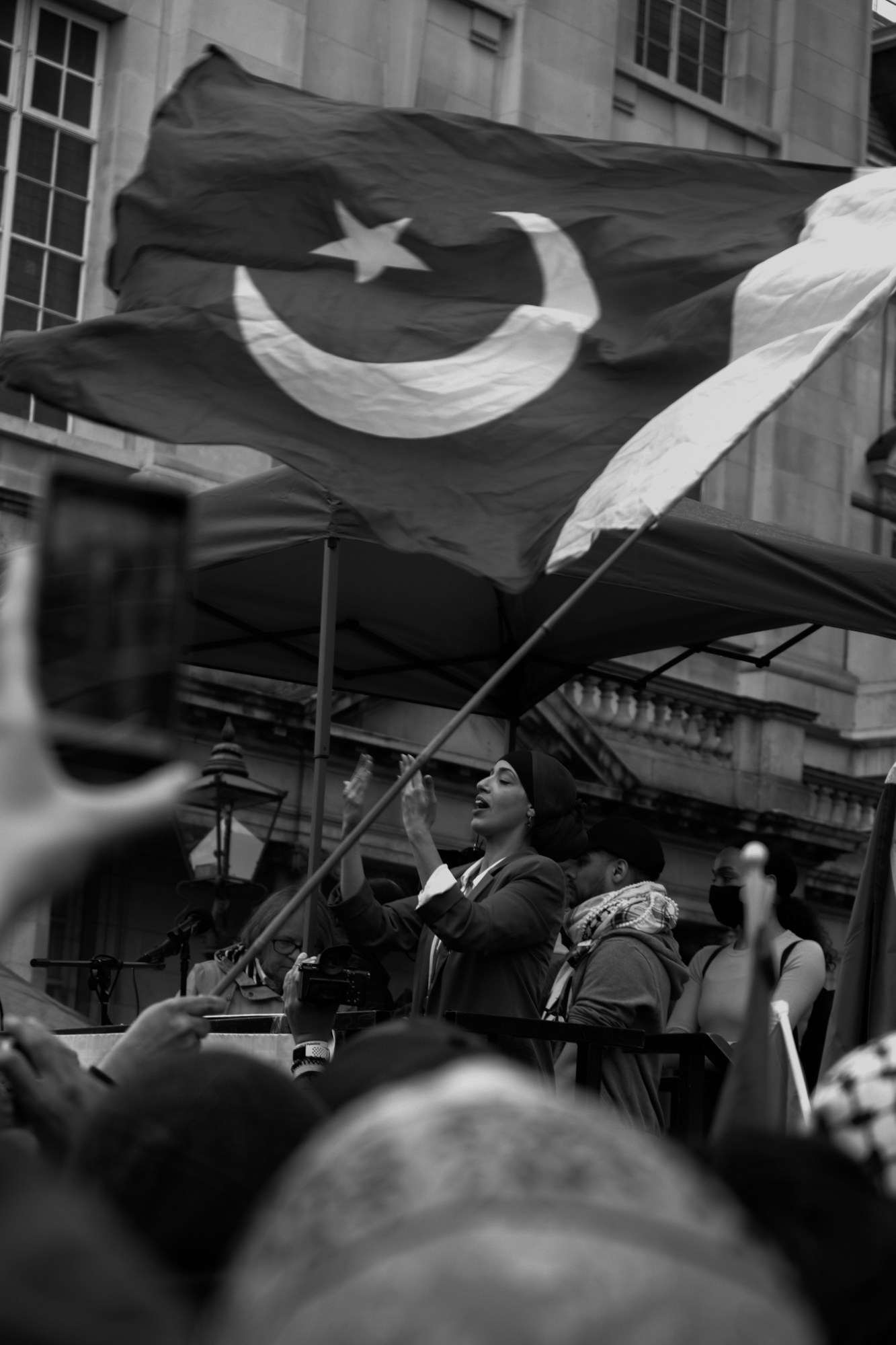 below a waving flag, a woman in a headscarf stands with her arms raised, shouting