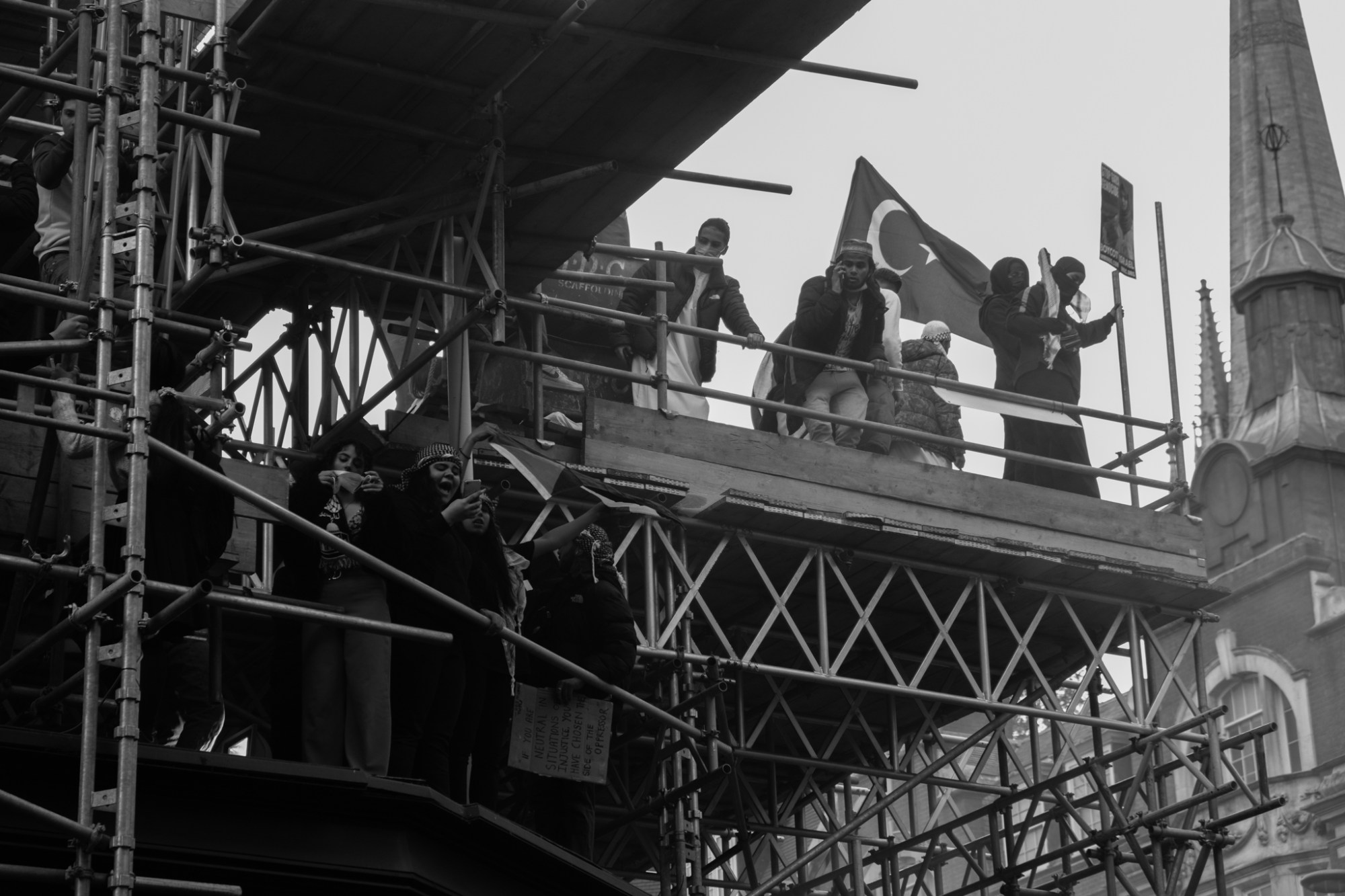 a group of young people stand on scaffolding with flags, banners and phones raised