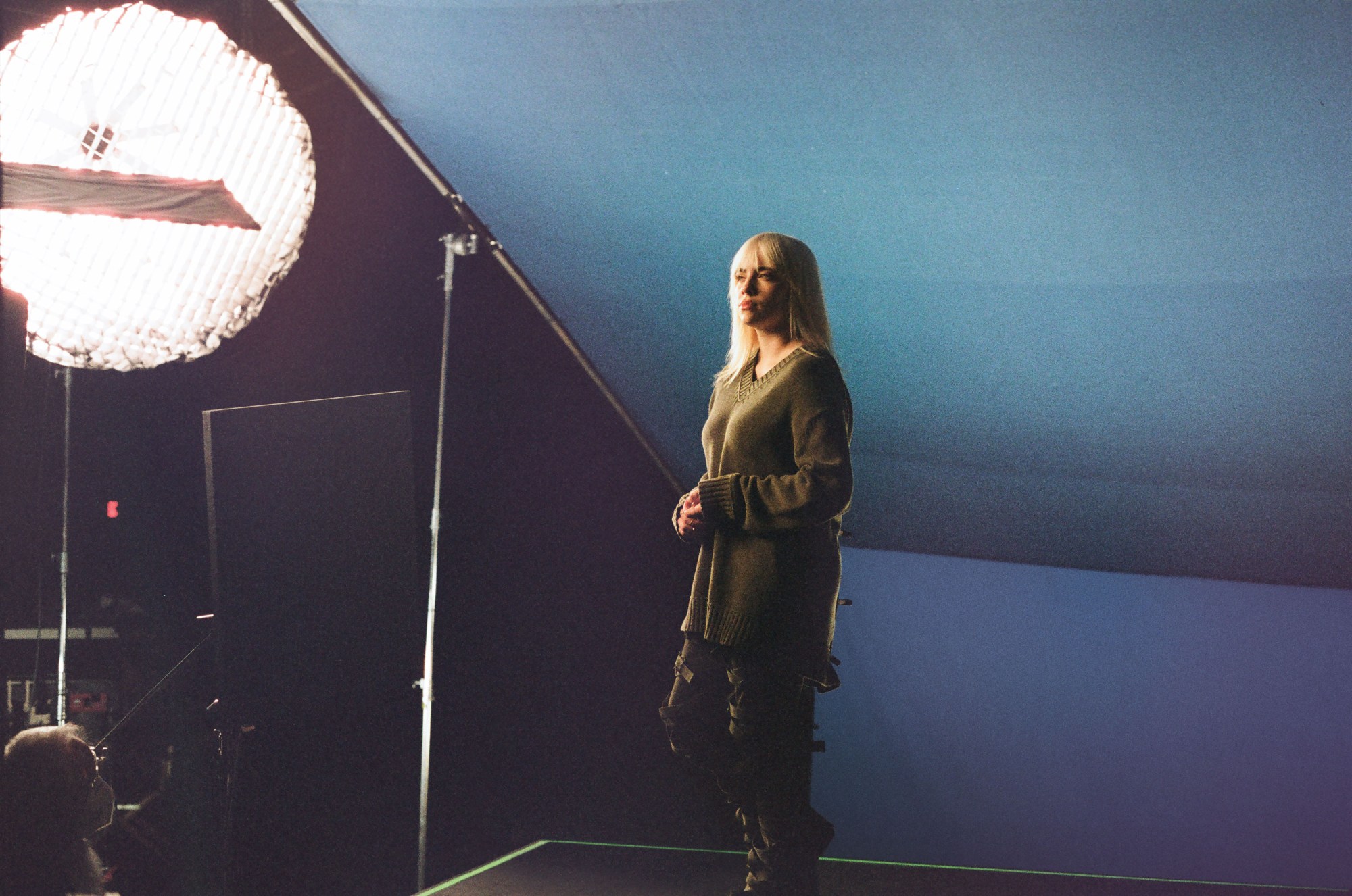 Billie Eilish with blonde hair in a green sweater, standing in front of a blue backdrop