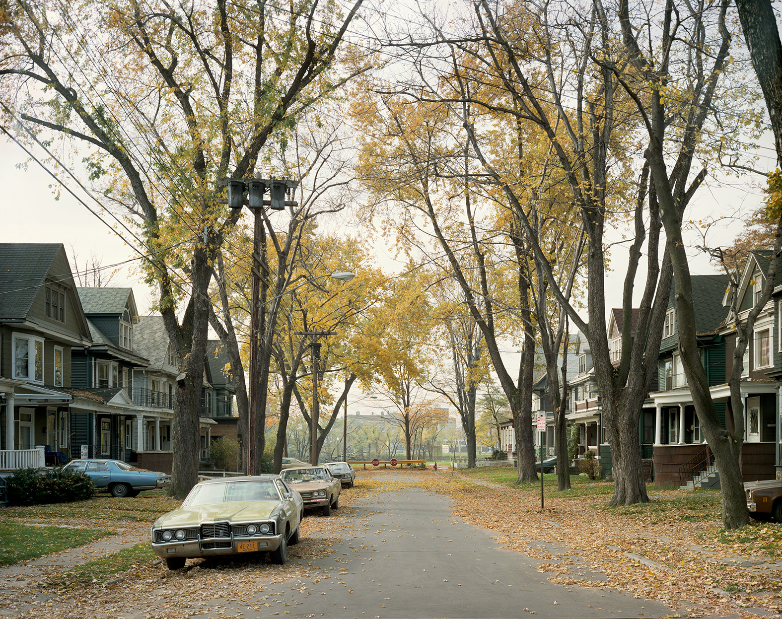 An empty residential street with parked cars and fallen leaves, 1977.