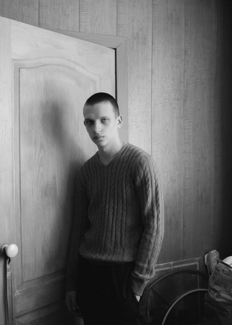 a young siberian man in a v-neck sweater rests against a wood panelled wall