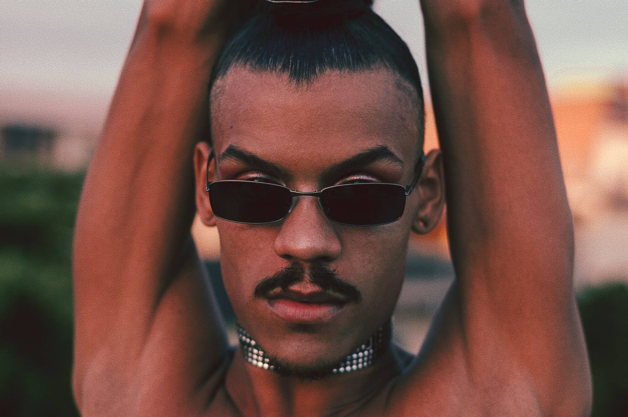 a man with tiny sunglasses and a chainmail choker on with his arms up