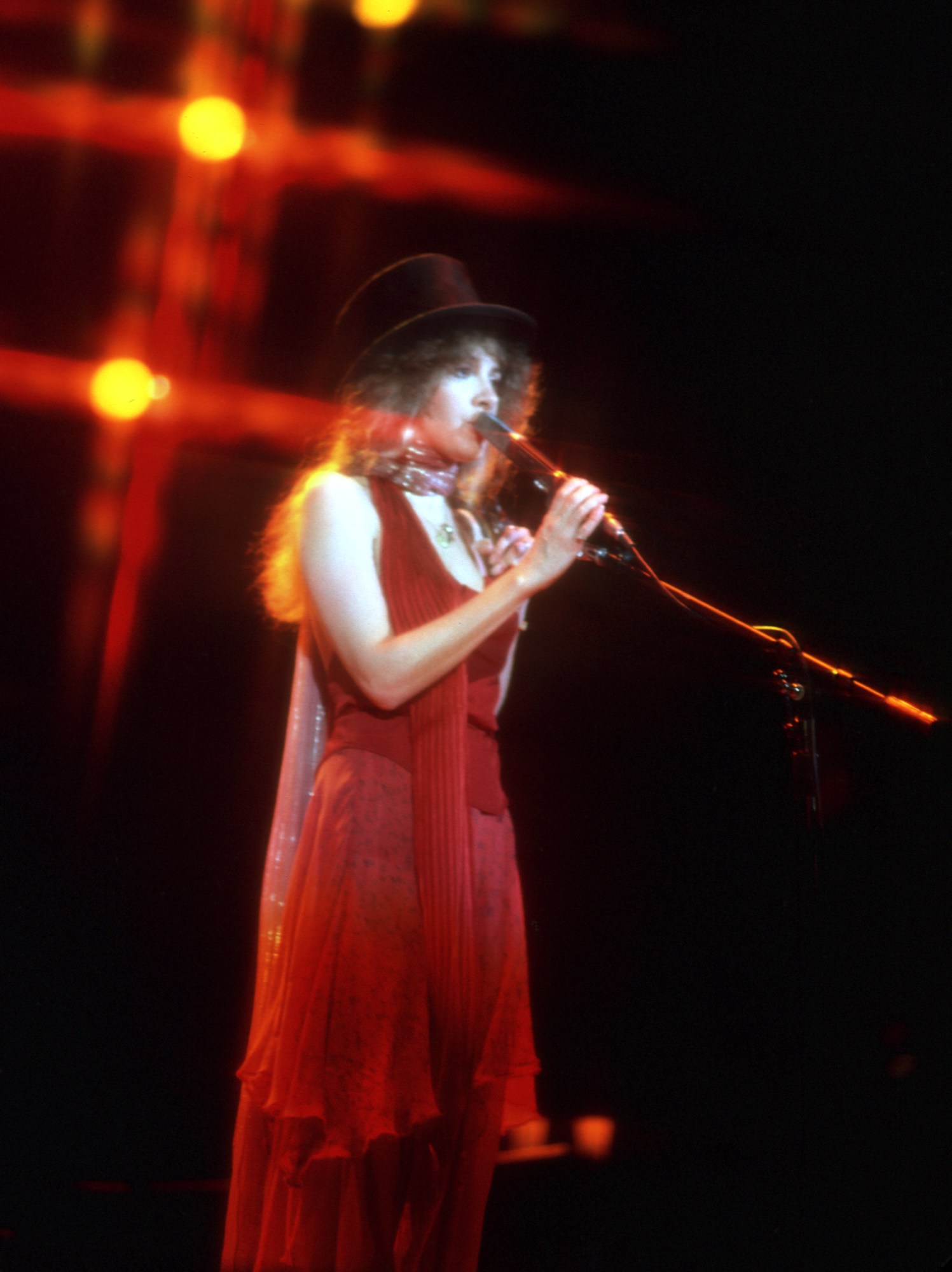 stevie nicks wearing a flowing red chiffon dress and black top hat while singing