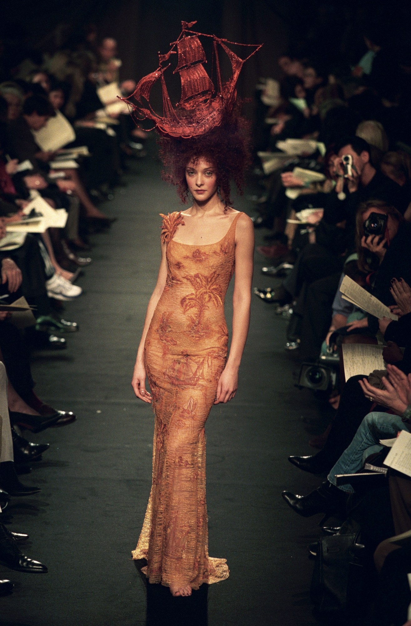 A model stands on a runway, wearing a Jean Paul Gaultier couture gown with a ship headpiece