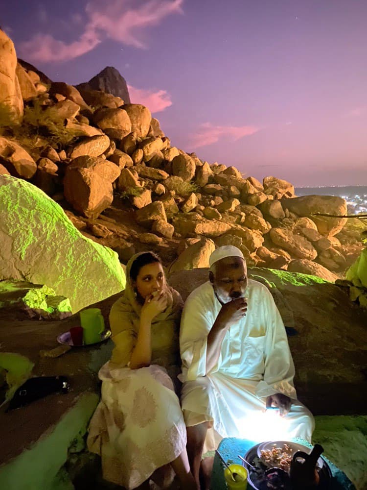 a couple sitting eating lunch on a mountain surrounded by orange rocks and a purple sky.