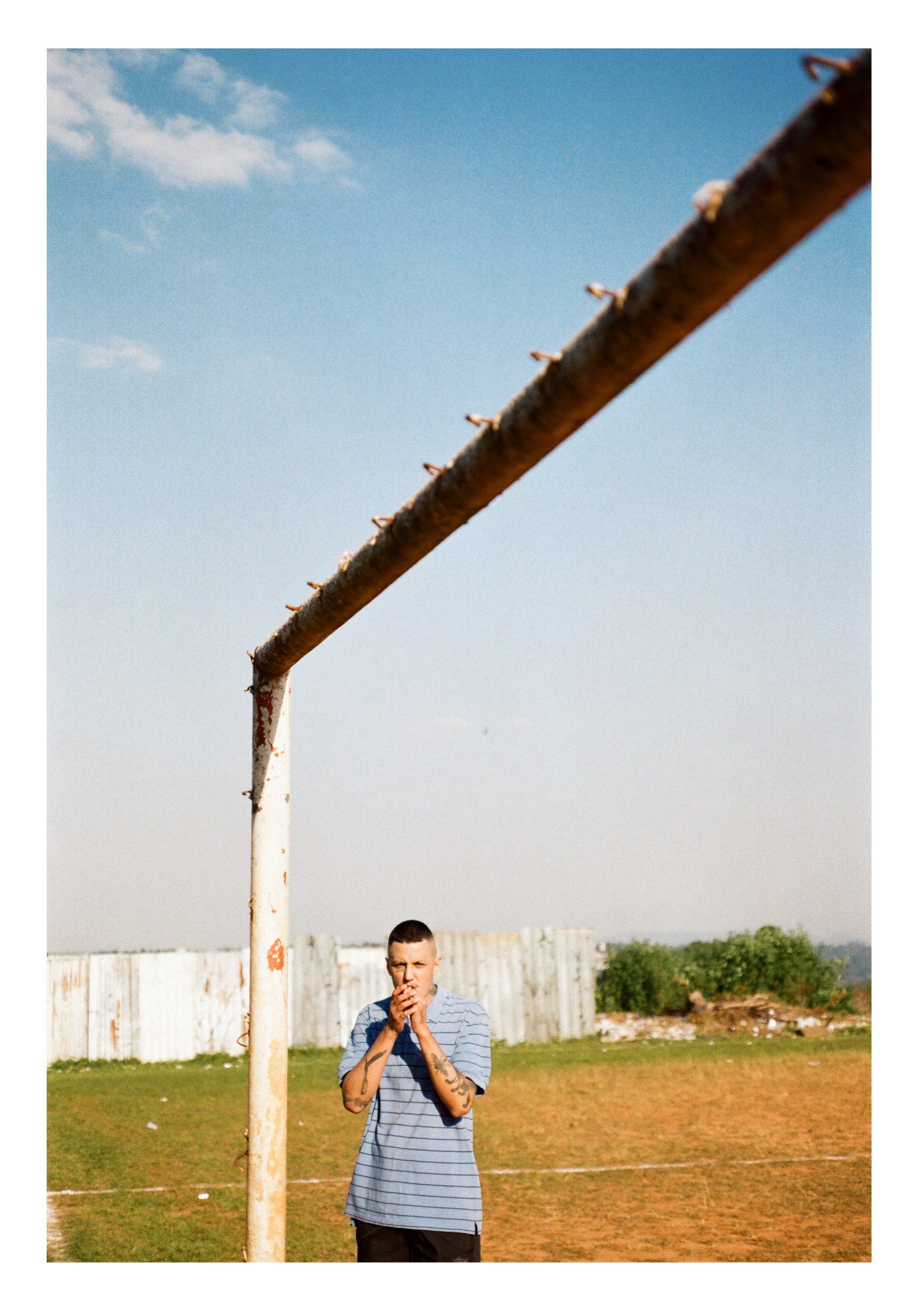 A man in a striped blue t-shirt standing under a rusted football goal post on a pitch in the sun.