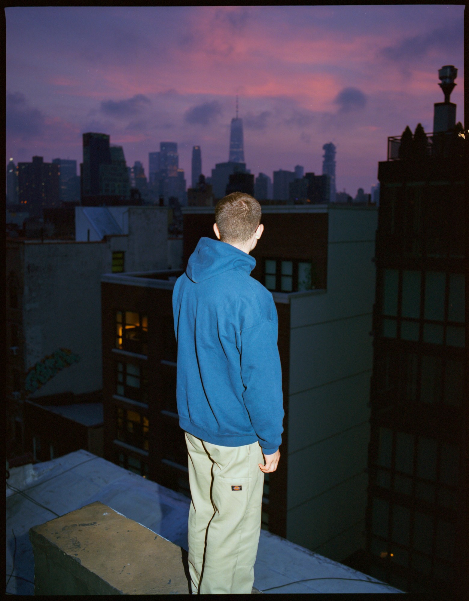 Man in blue hoodie standing on a roof over the purple New York sunset