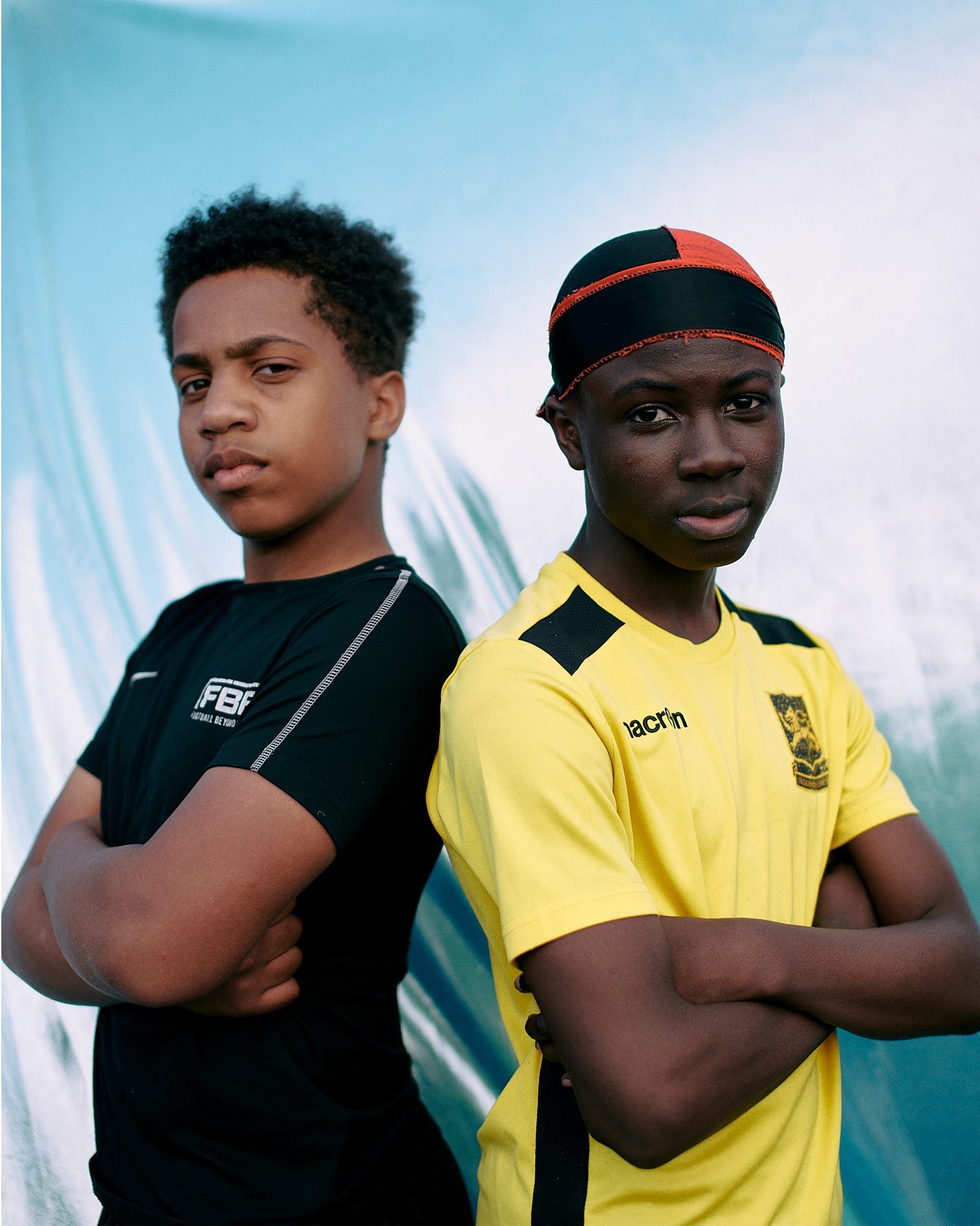 Two boys pose back to back with arms folded against a blue background