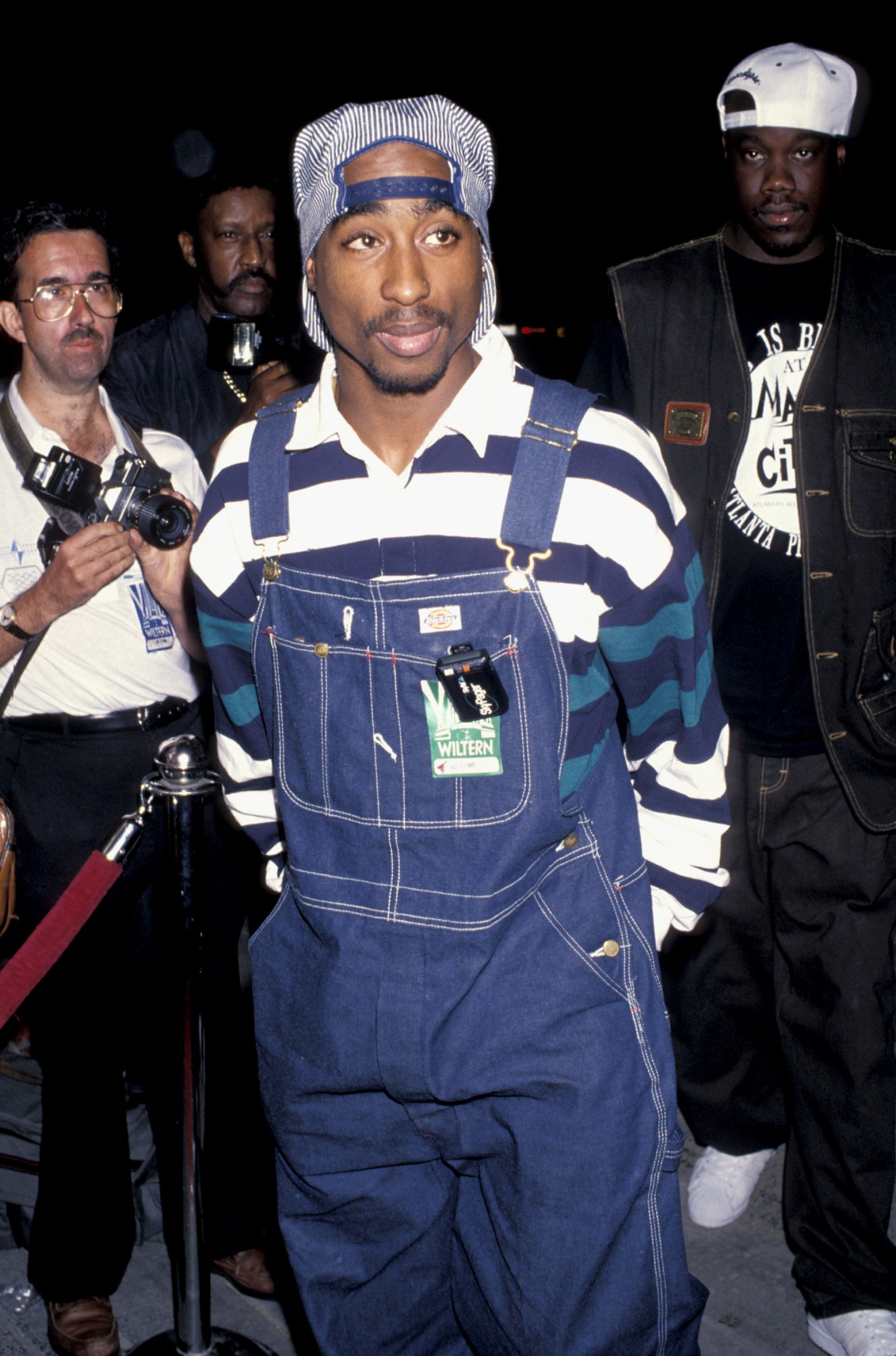 tupac with his hands in the pockets of his overalls and a hat on