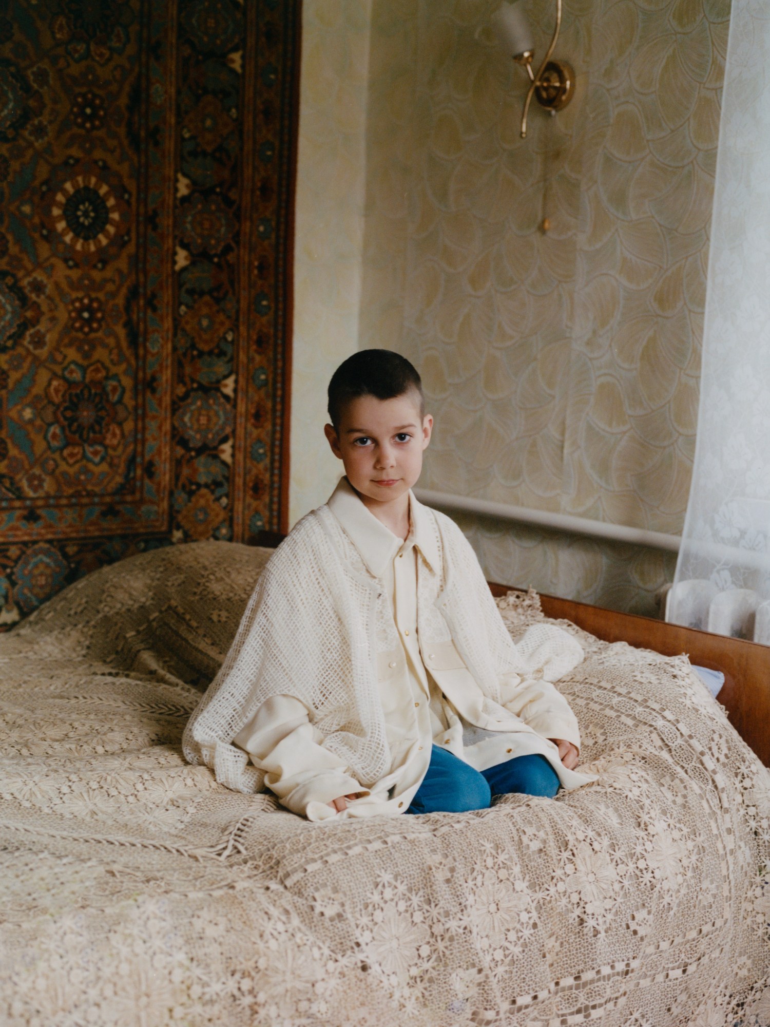 a young boy in a large cardigan sits on a bed