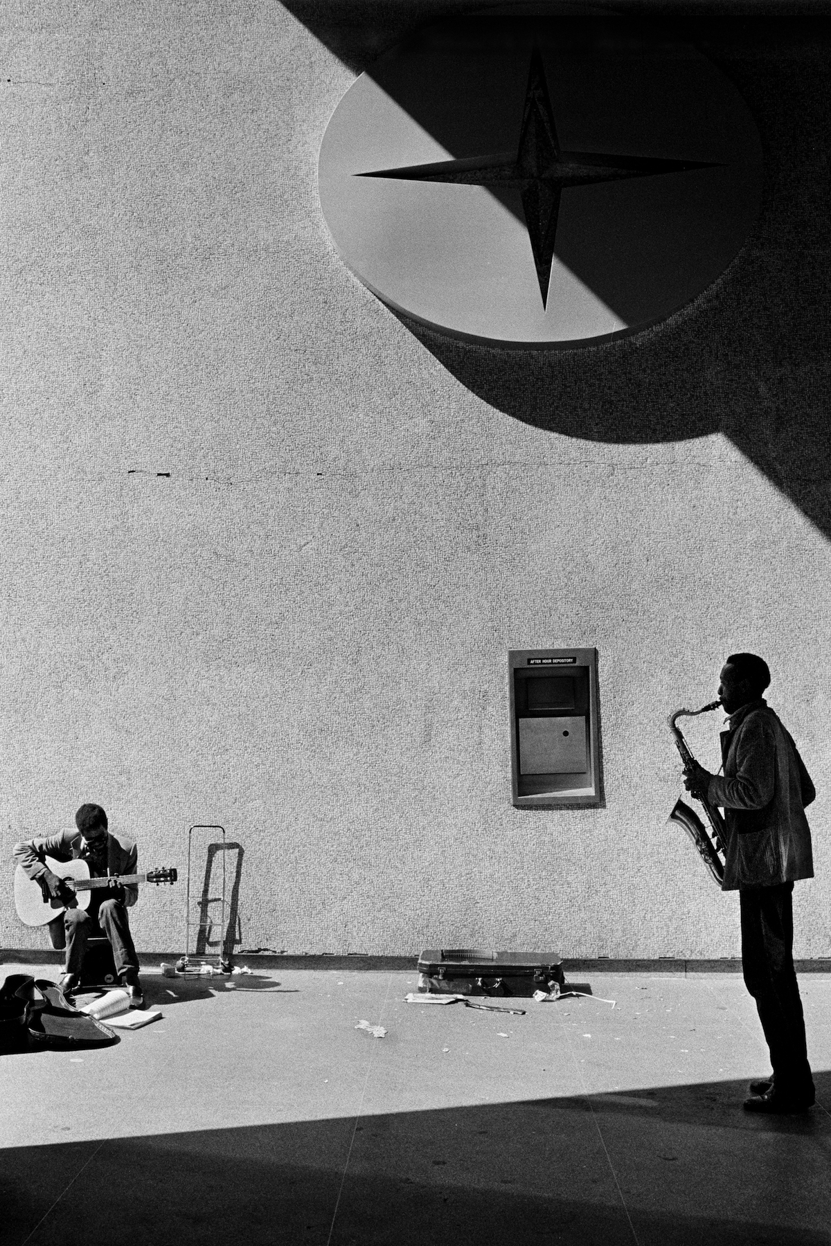 two men, one playing a guitar in the sun, the other playing a saxophone in the shade