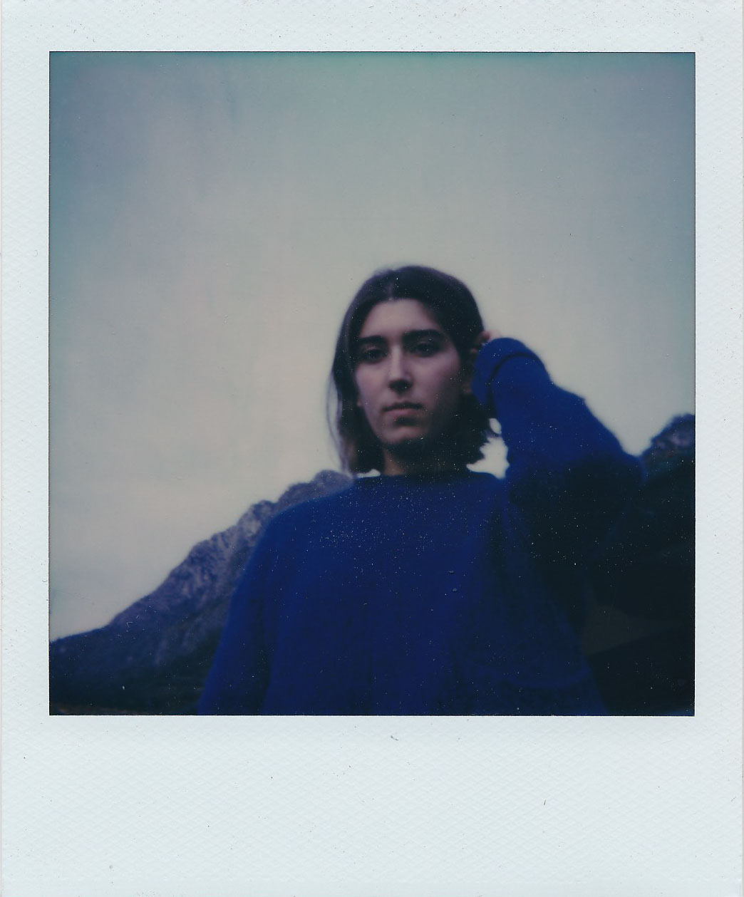 a woman in a blue jumper tucks her hair behind her ear in front of a mountain