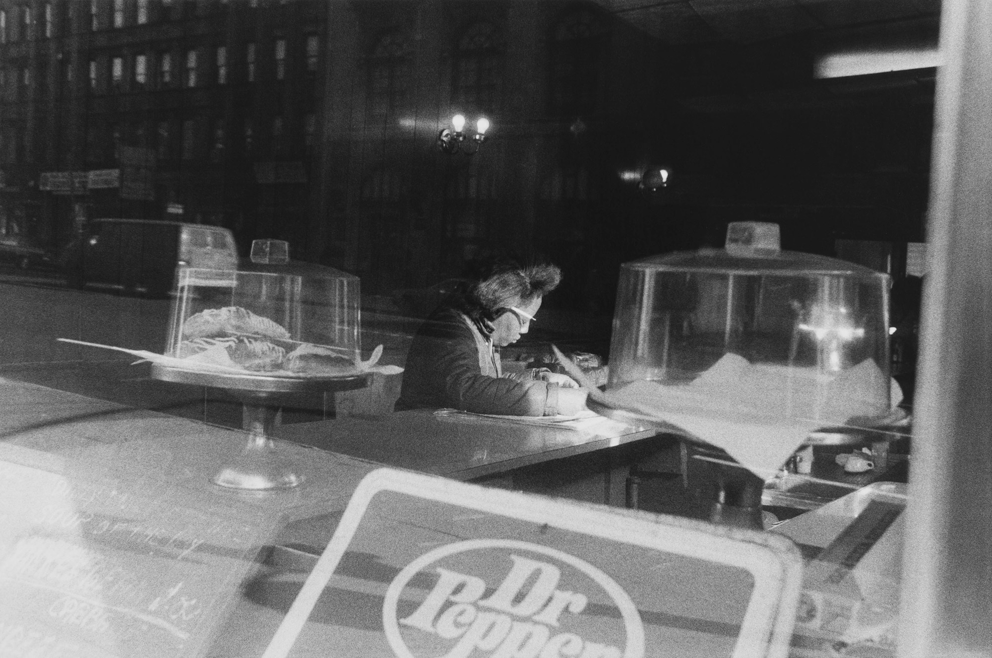 a woman sits at the counter of a cafe reading something, a dr pepper sign can be seen in the foreground