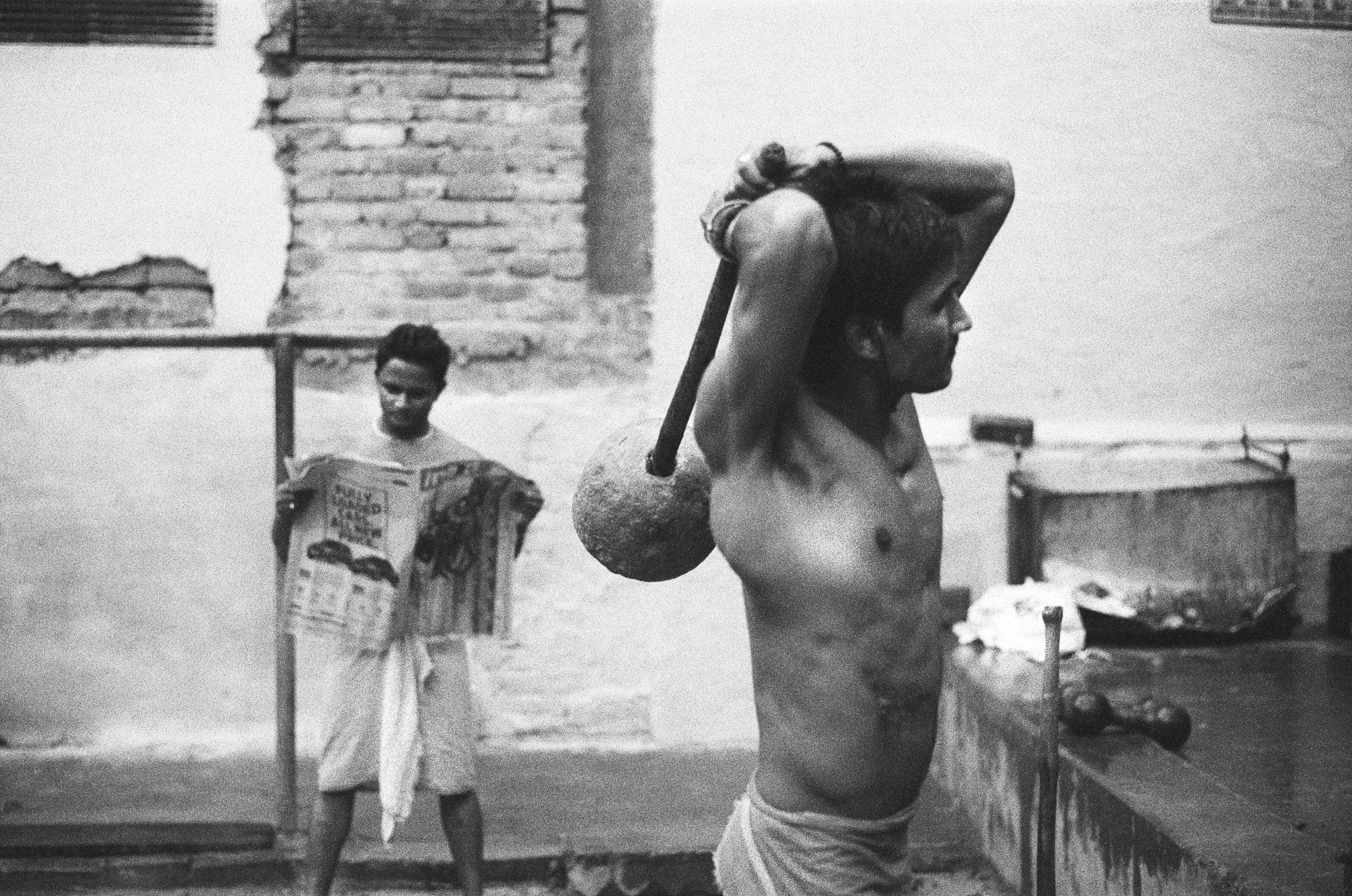 a man trains with a handmade weight while another reads a newspaper