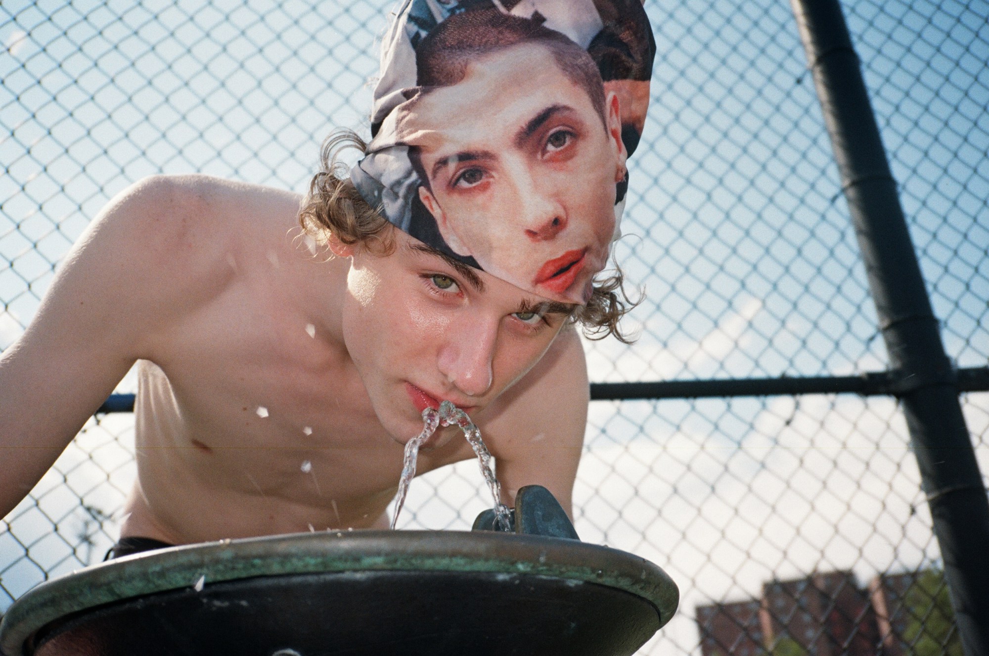 a man wearing a headscarf with a face on it and no top drinks from a water fountain