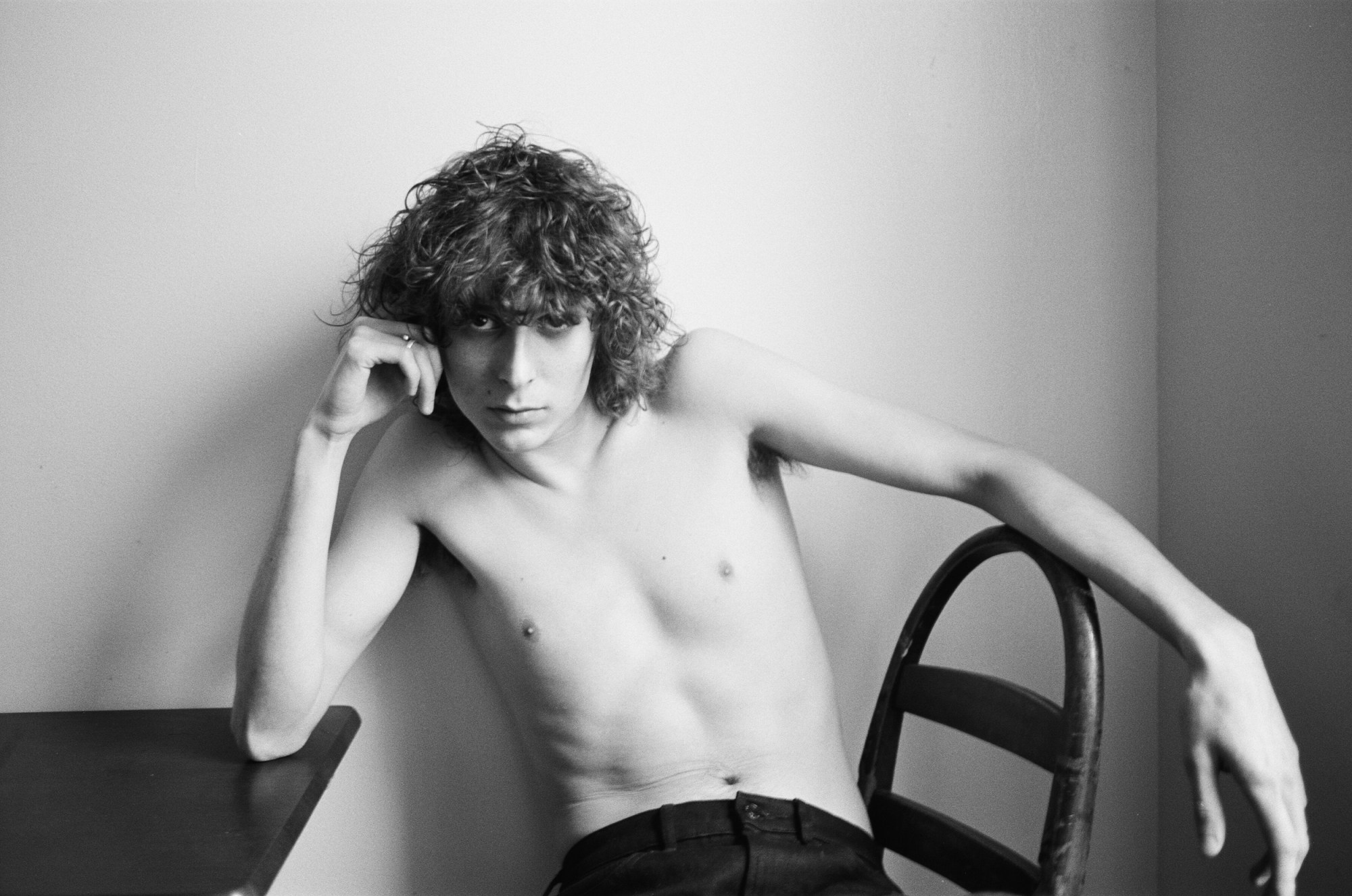 a topless man with curly hair sits on a chair