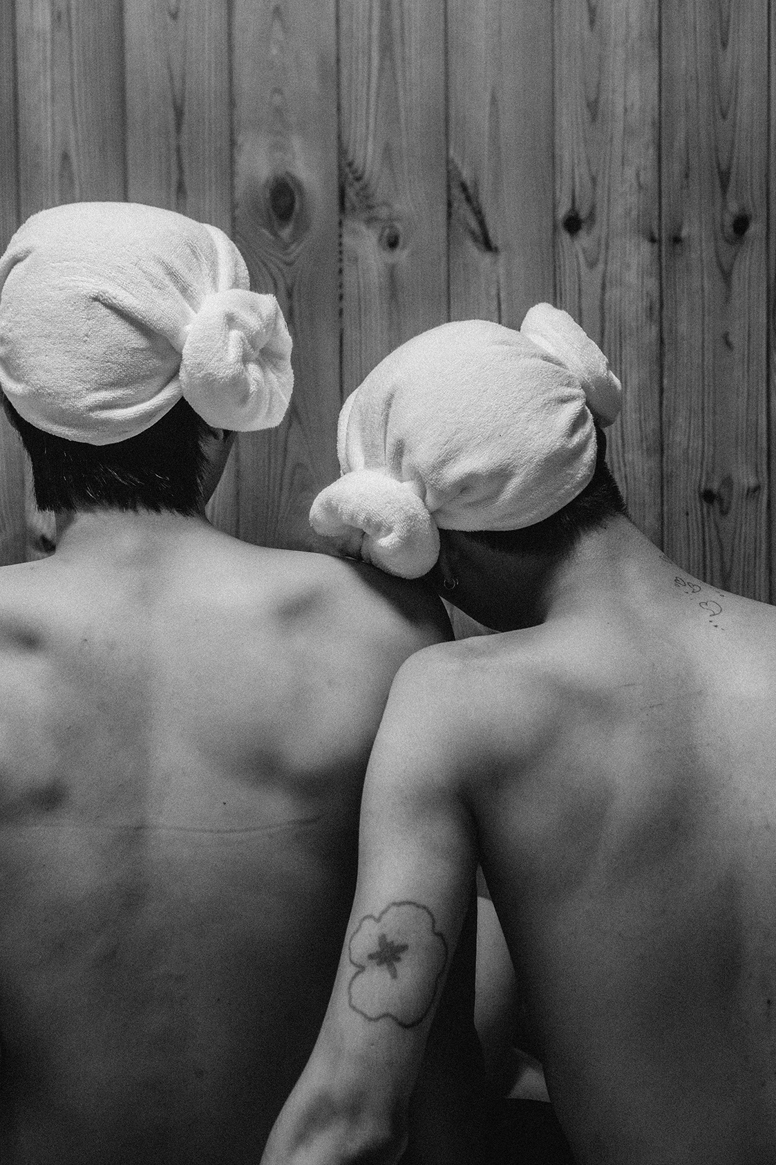 the naked backs of two men, one leaning his head on the other's shoulder