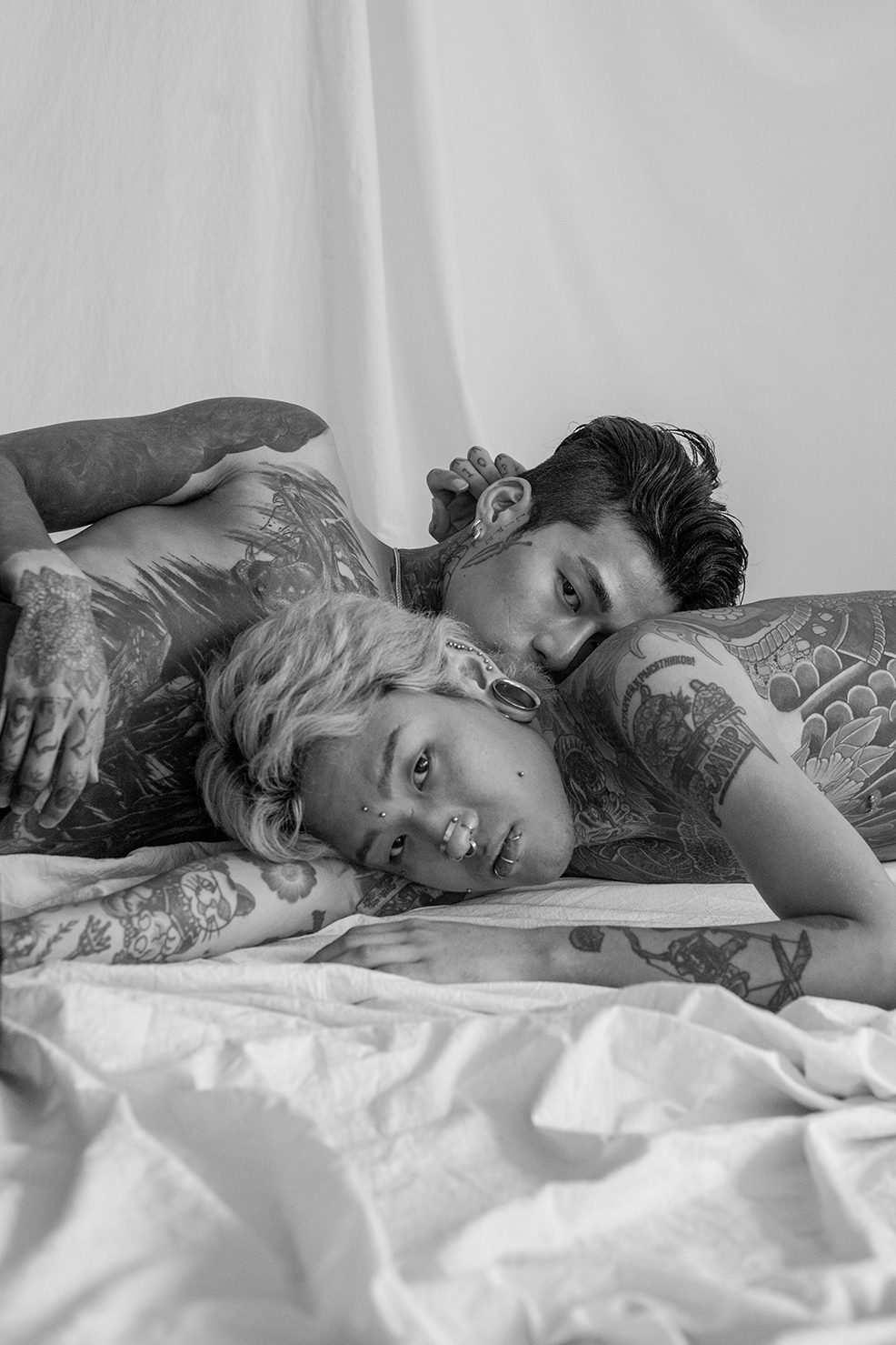 two heavily tattooed men, one with bleached hair and piercings, lie on a bed