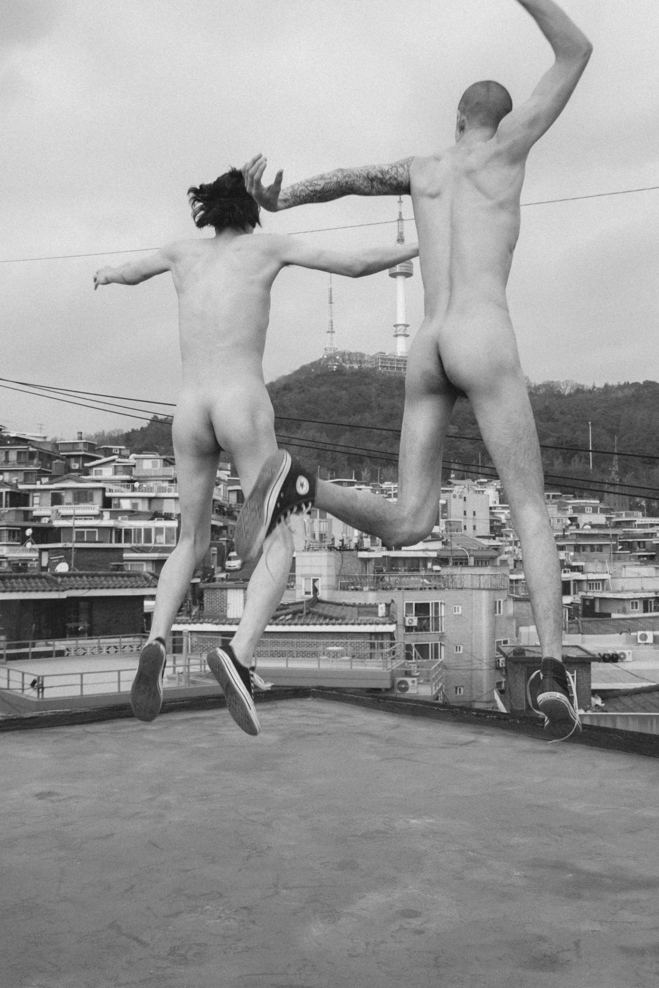 two naked men wearing converse jump up into the air on a rooftop