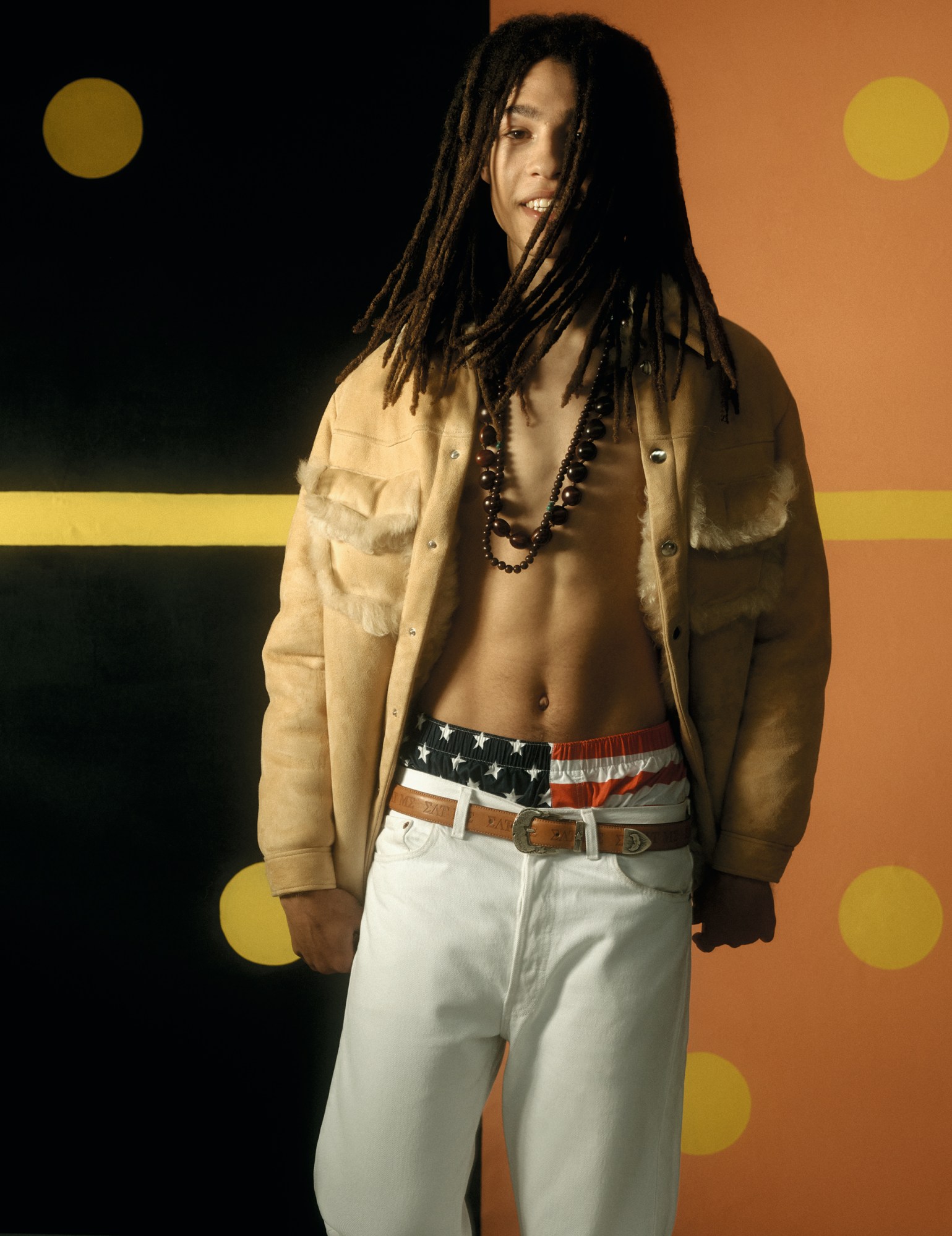 a model wearing white jeans with american flag boxers showing, with an open beige suede jacket and beads