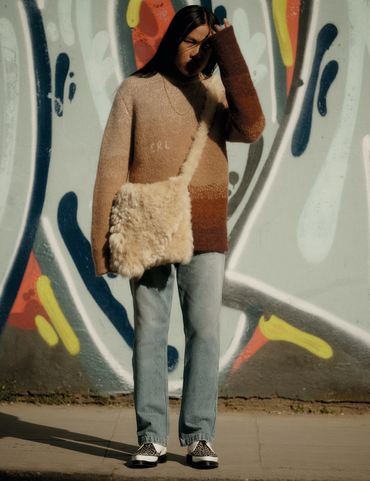 a model standing against a graffiti wall wearing a sweater, jeans and a fluffy cross-body bag