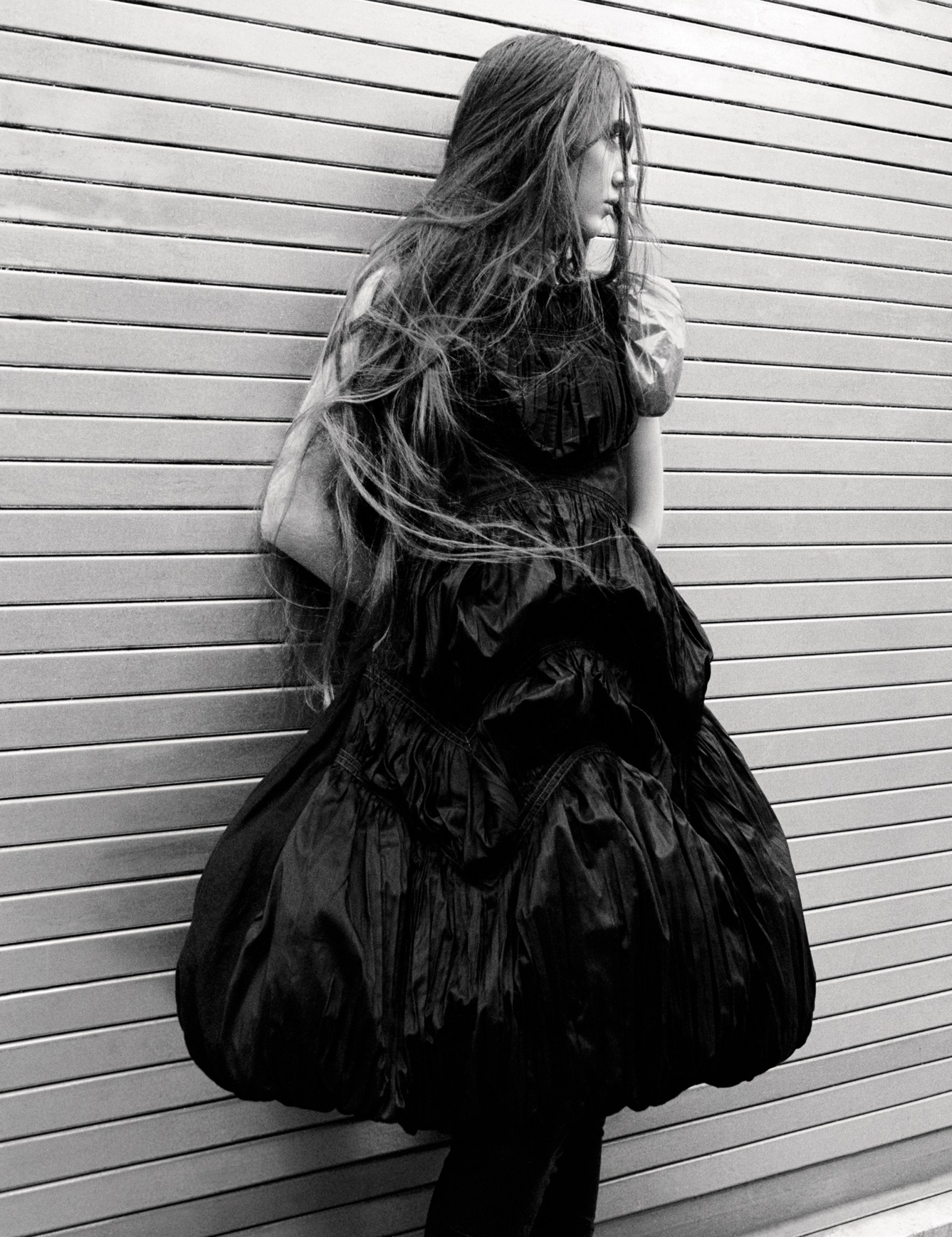 a model with long messy hair in a black poofy dress, stands with their back to a wall