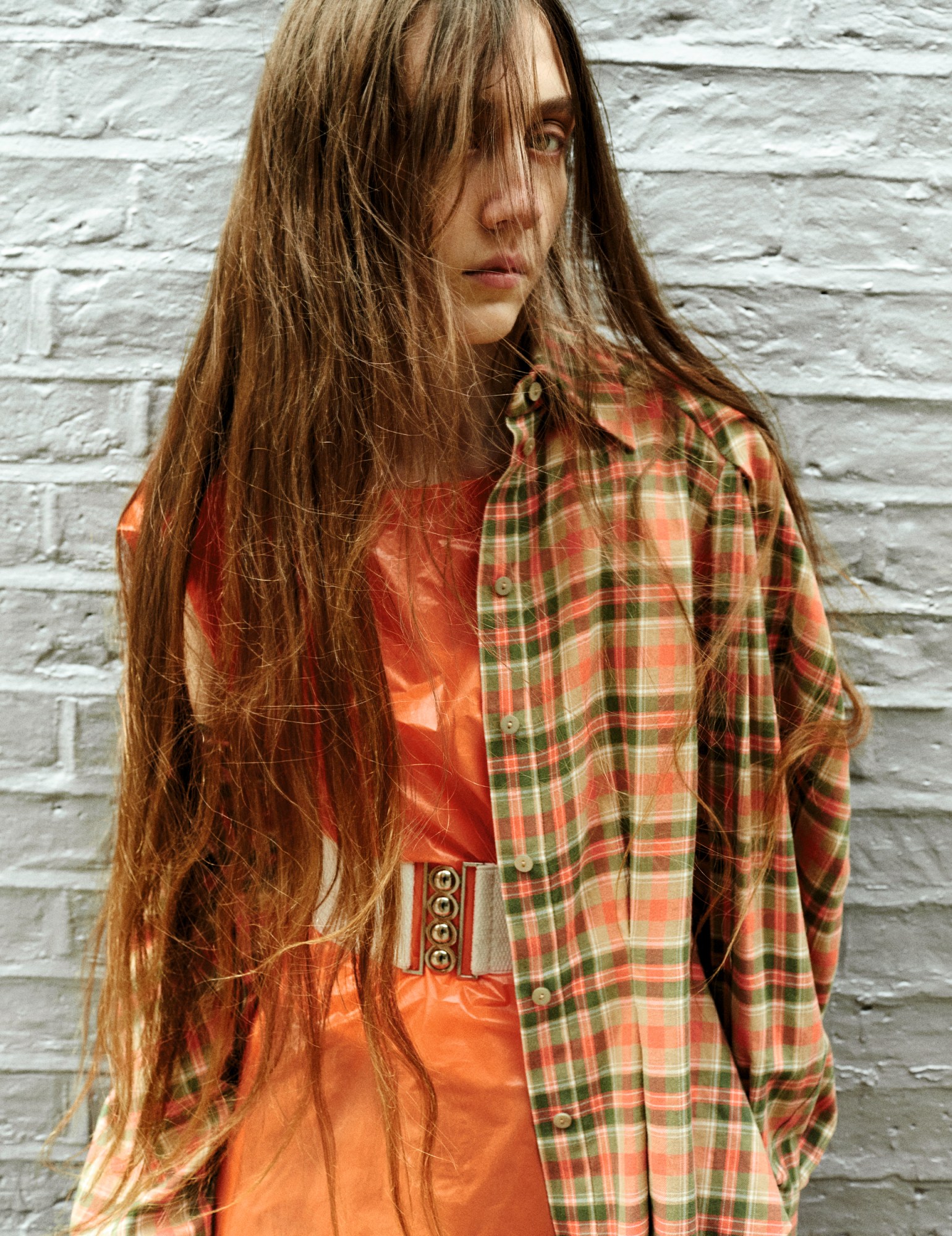 a model looks to the camera from behind their long brown hair, wearing a checked shirt over a belted orange dress