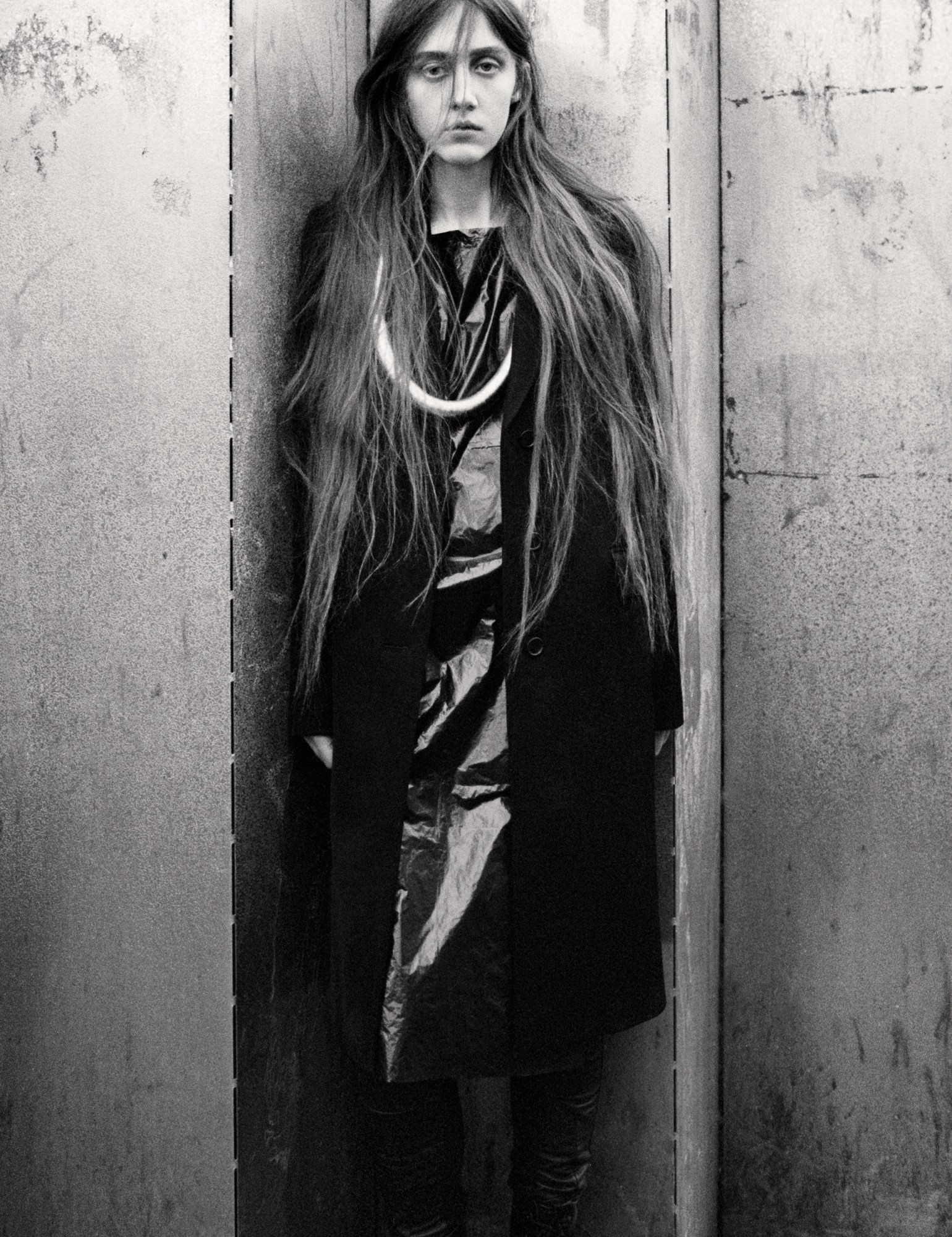 a model with long hair stands against a zig-zag panelled wall in a long overcoat