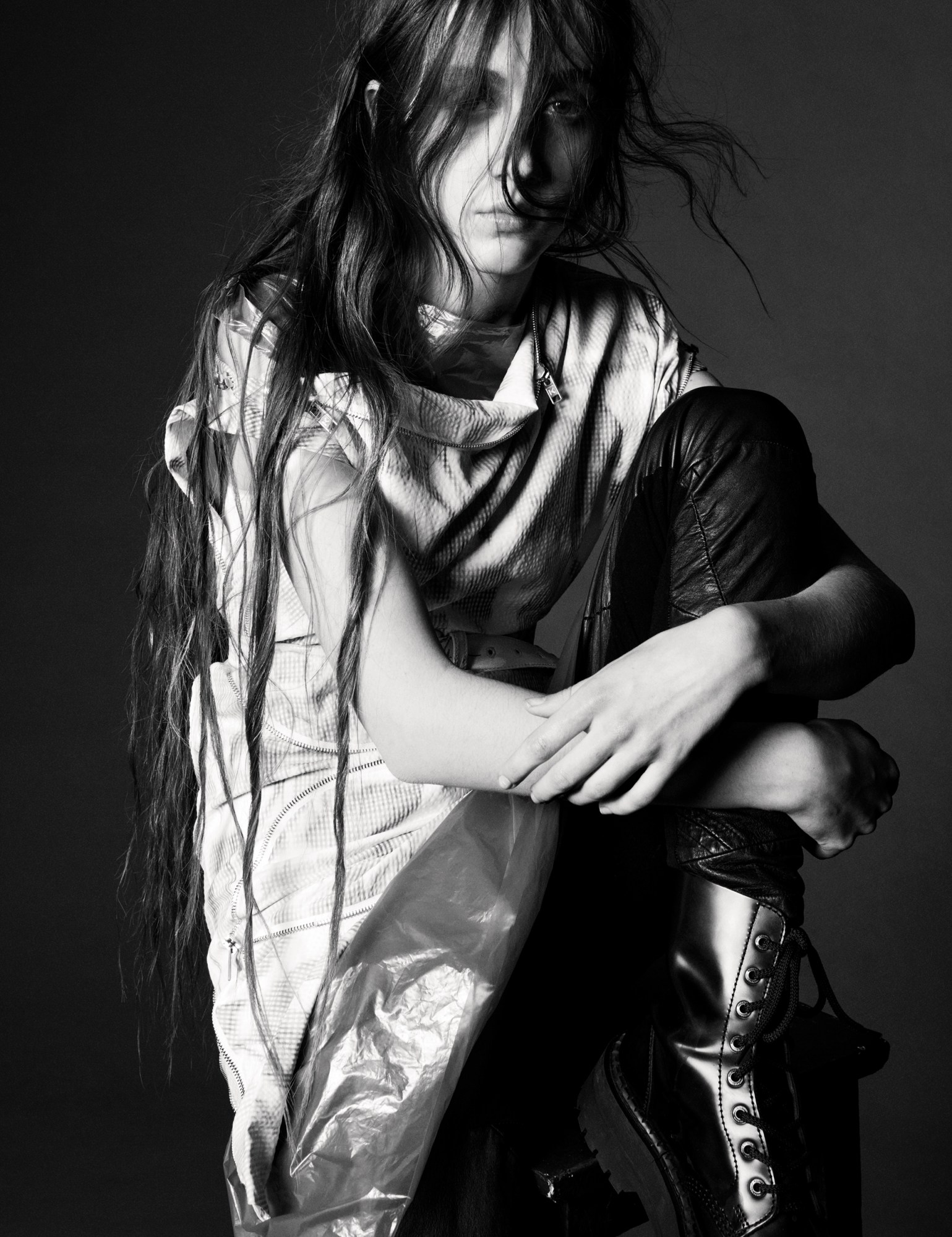 a model sits on a stool, cradling one leg up to their body, looking to camera from behind their long messy hair