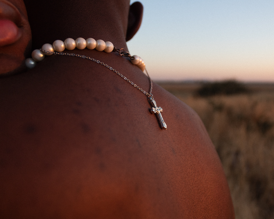 A close up of the top of a man's back with a pearl necklace around his neck and a crucifix chain dangling down his back. In the background is a grassy outdoor sunrise.