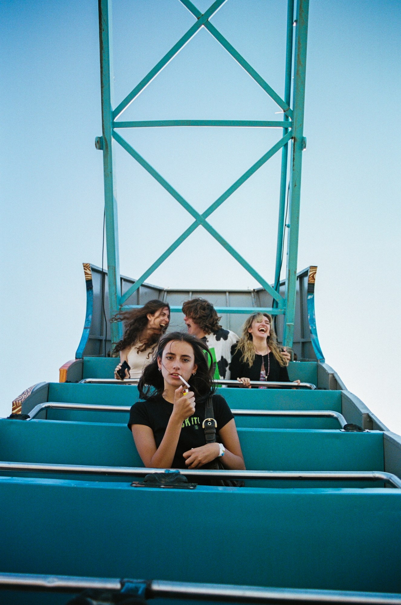 A girl lights a cigarette calmly as she sits alone on a rollercoaster car and looks directly into the camera. Behind her is three girls screaming and laughing.