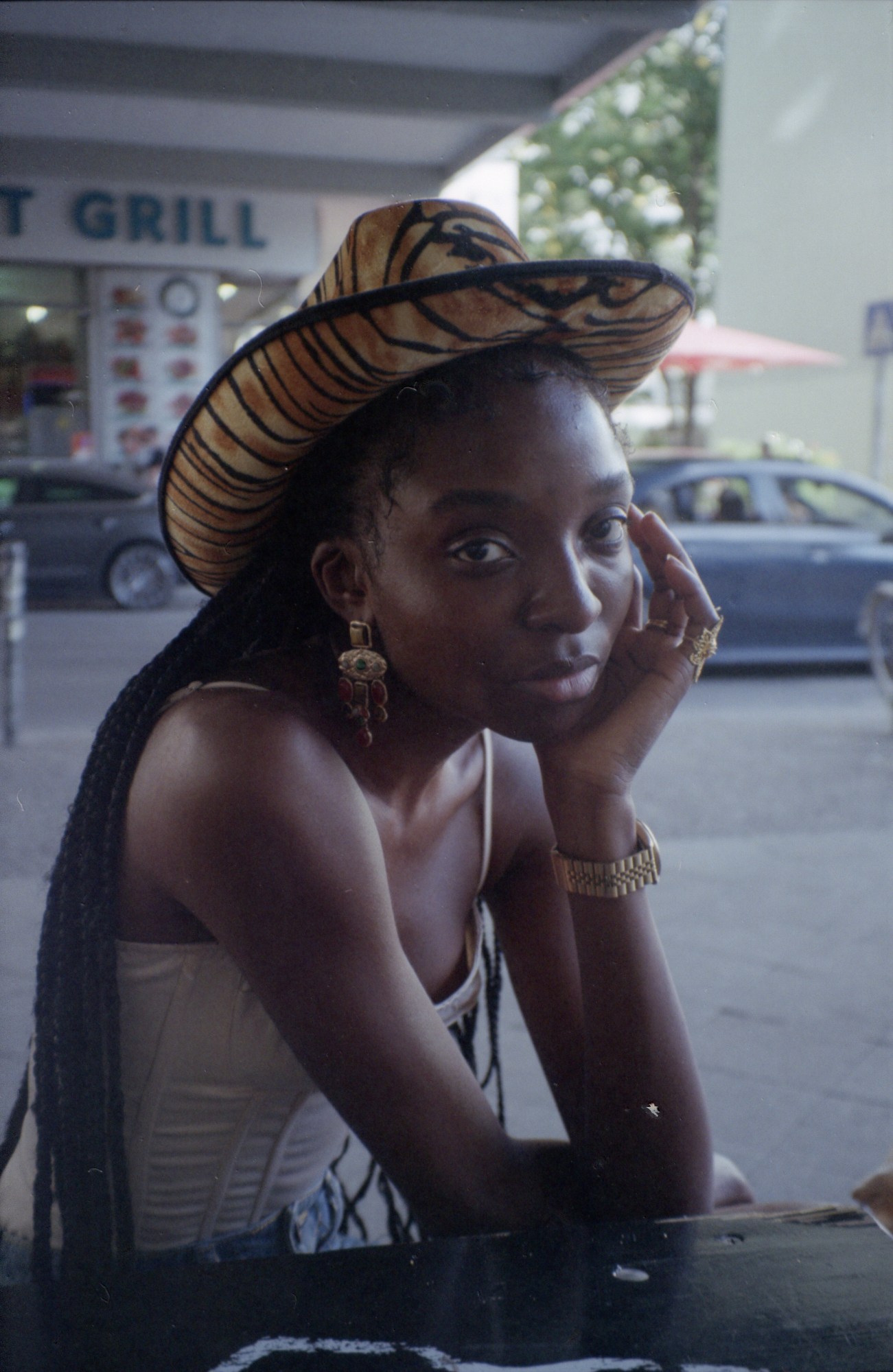 A girl sitting on a bench wearing a white corset top, blue jeans, gold watch and earrings and tiger print cowboy hat. her head rests in her hand while behind her is a corner shop and cars.