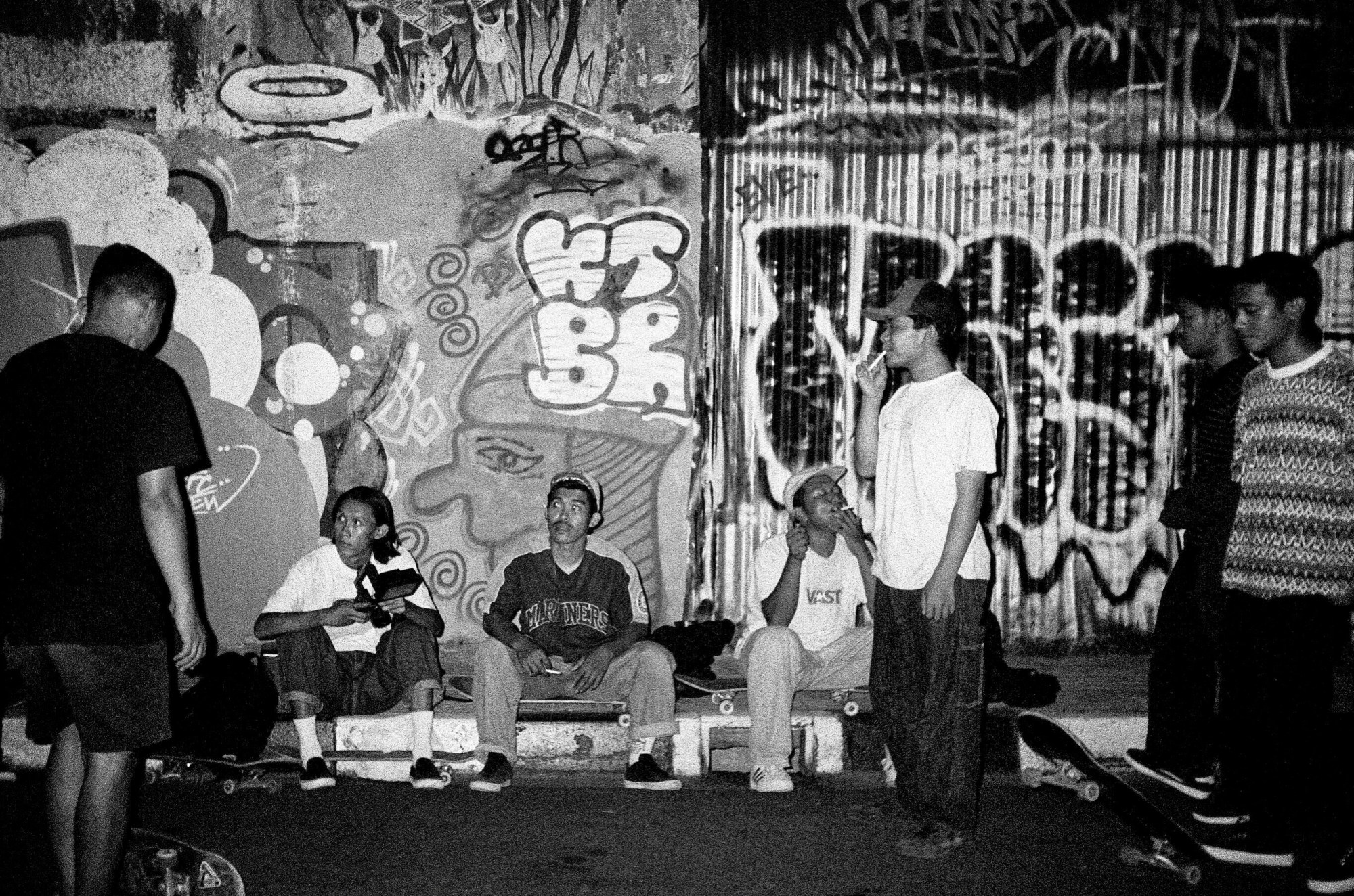 Skateboarders sitting on their boards and smoking outside a graffiti covered wall.