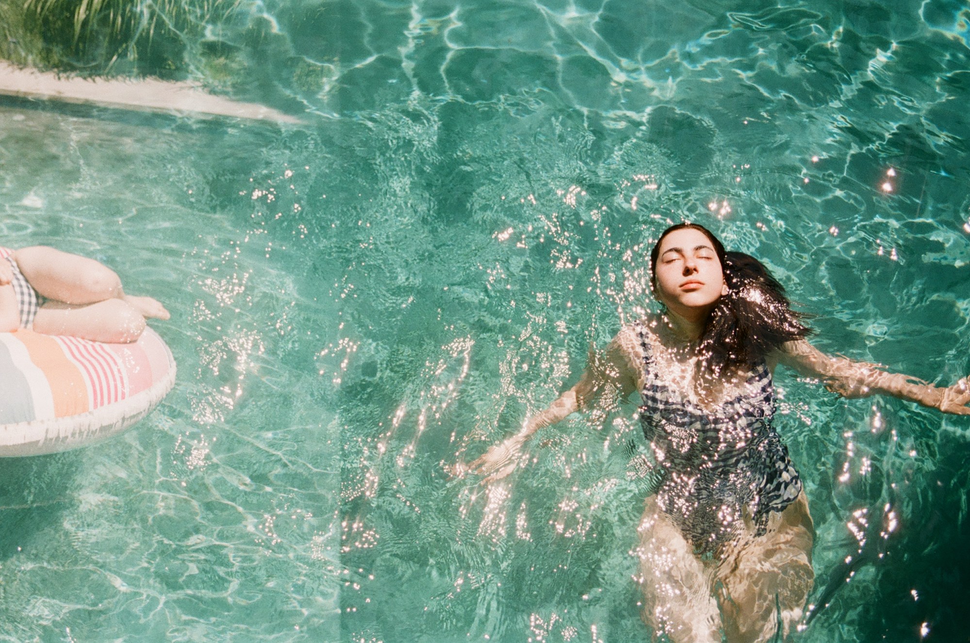 A woman lays back in a glimmering pool. On the side is grass and a child laying on an inflatable.