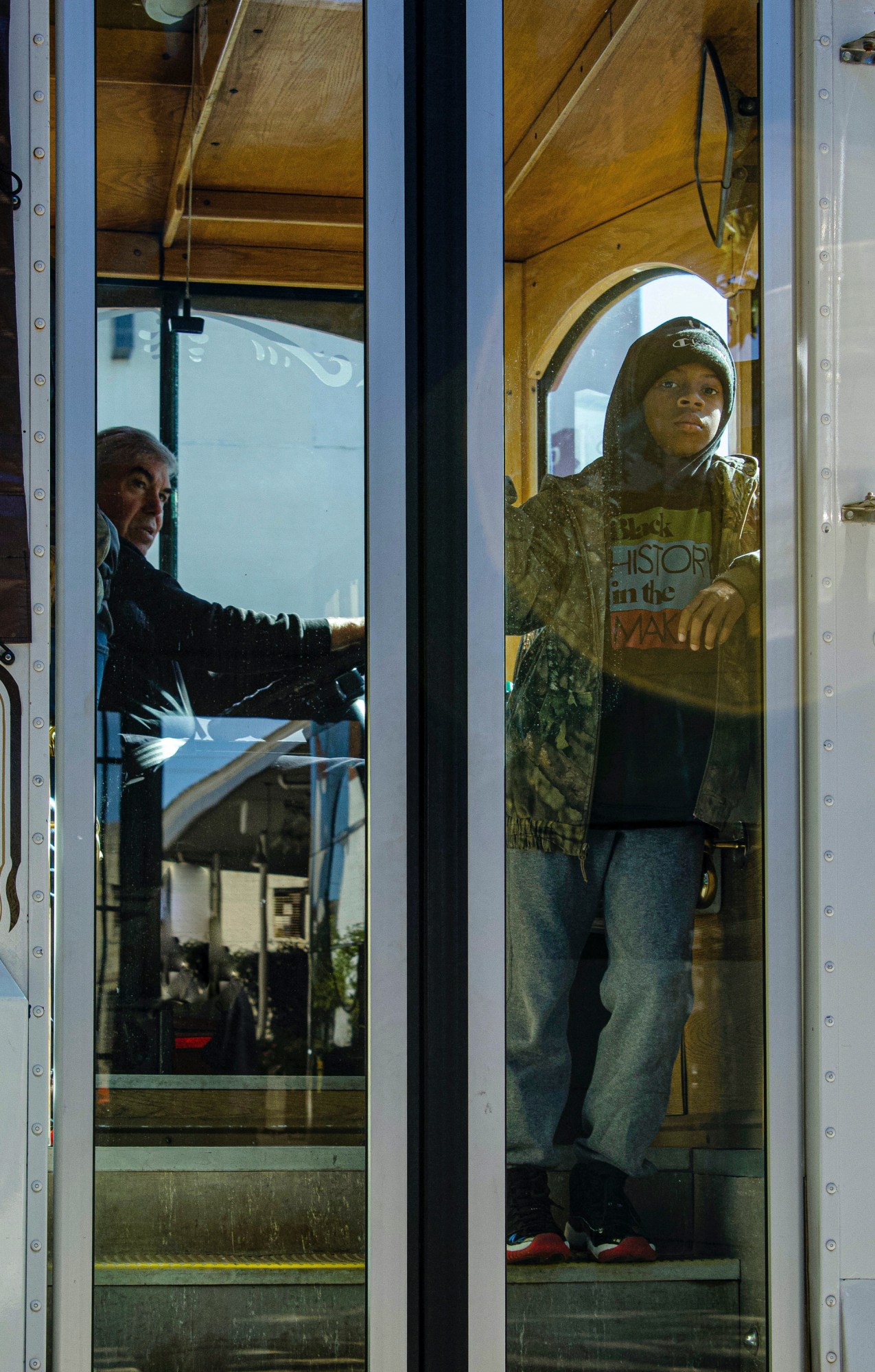 A little boy in jeans and a hoodie standing in the entrance of a bus looking out the door windows. The driver stares at him.