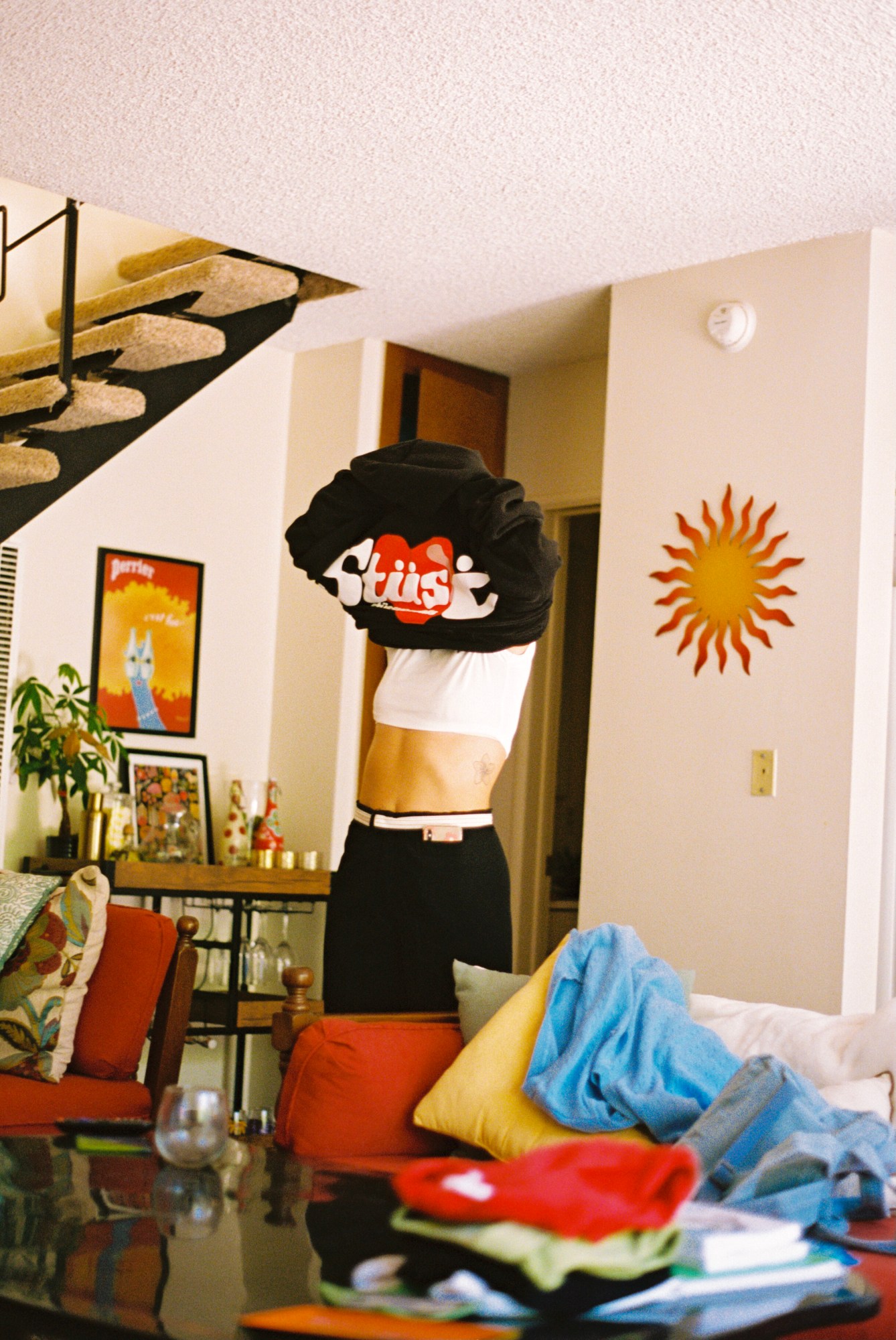 A woman lifts a top over her head as she stands in a living room by the stares. A sun and cat paintings are on the wall and the sofas have clothes and cushions on them.