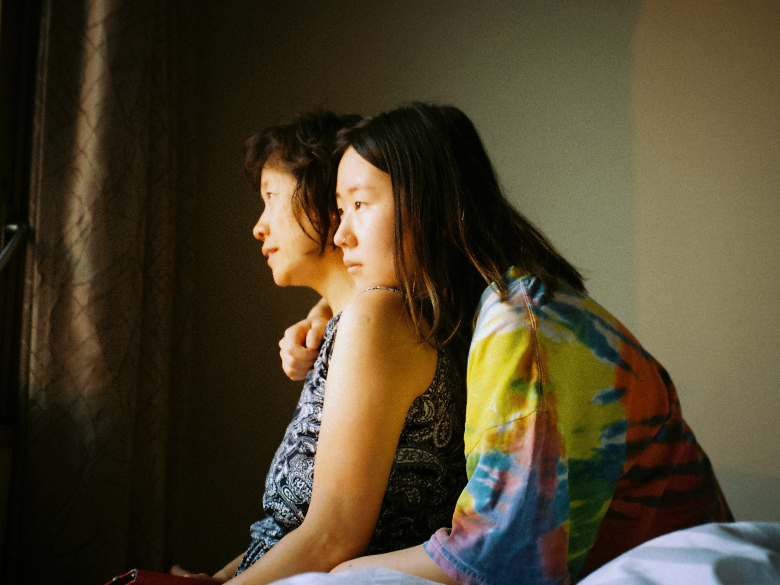 a young girl and her mum sit and look out the window. She wear a tie-dye top and her mum wears a black patterned blouse.