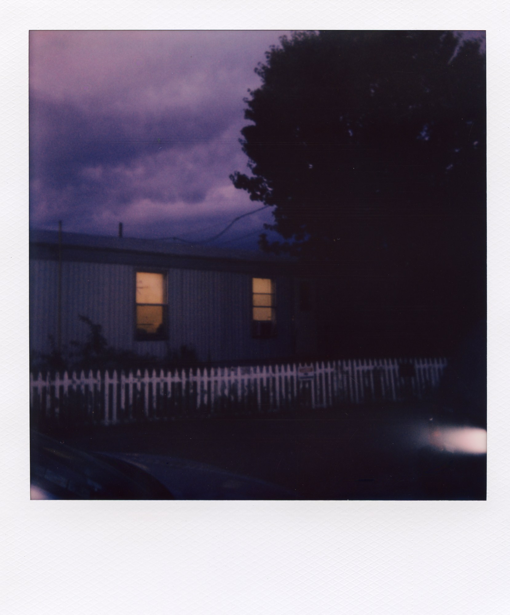 A bungalow with the lights on in the windows during a cloudy night. A white fence sits in front and a large tree is on the right.