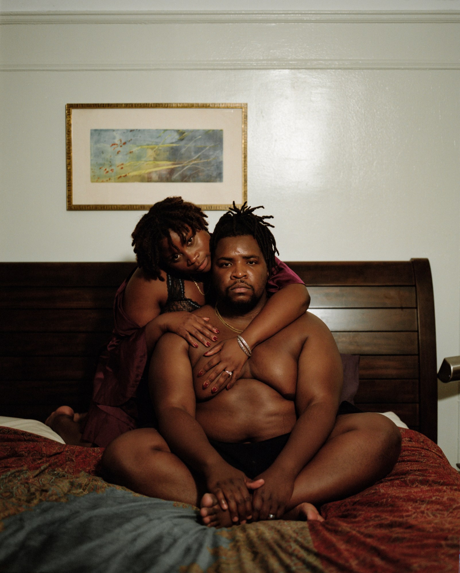 a topless man sits on a bed with a woman behind him wrapping her arms around him