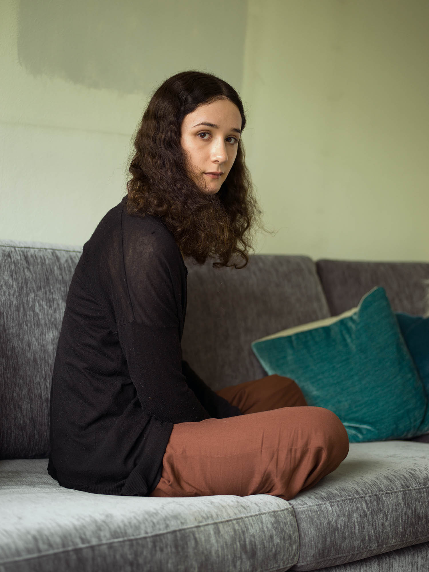 a woman sits on a sofa looking over her should at the camera