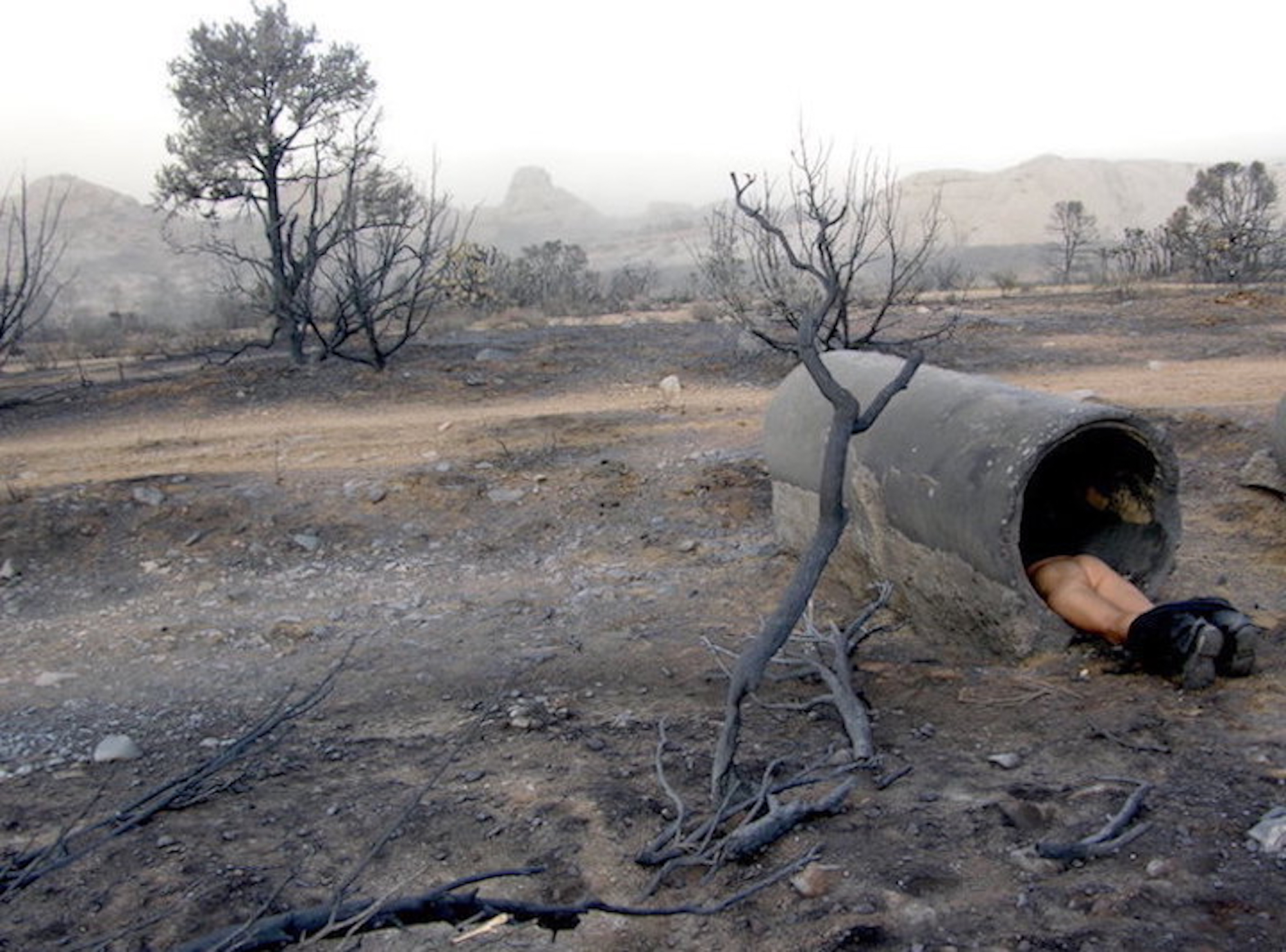 a burnt expanse of land with a naked man lying inside a concrete tube