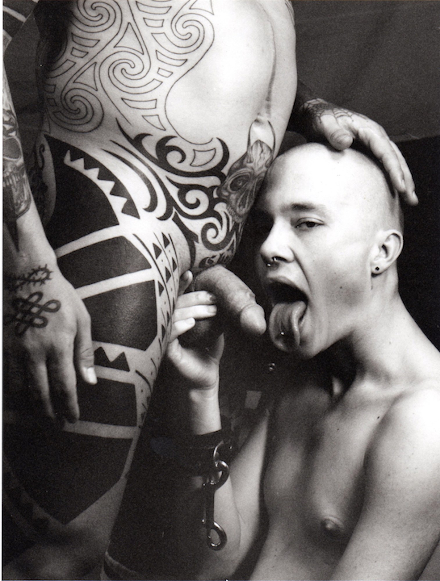 a man holds the penis of a heavily tattooed man next to his tongue