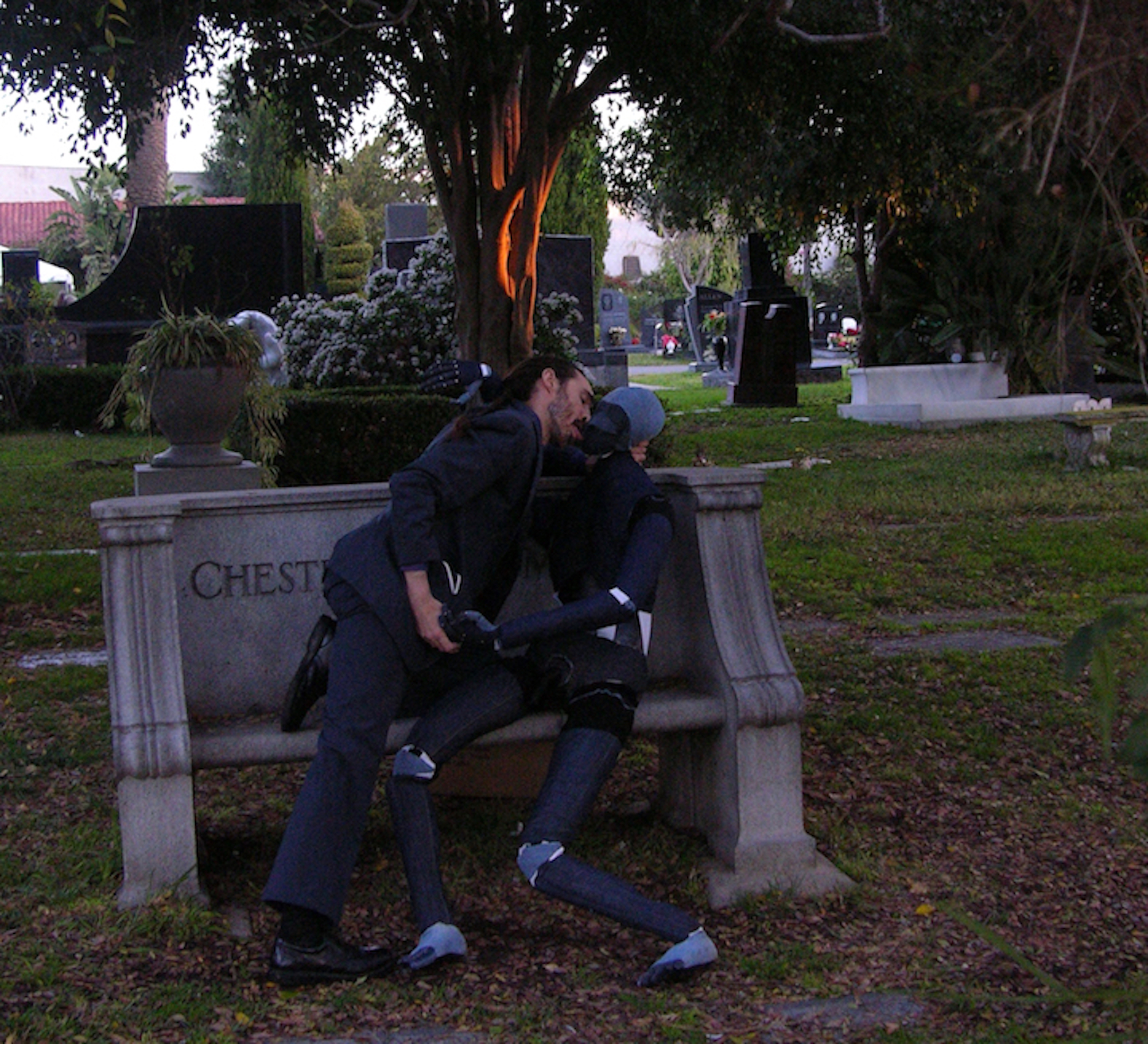 a man and a makeshift person kiss on a stone bench at a graveyard